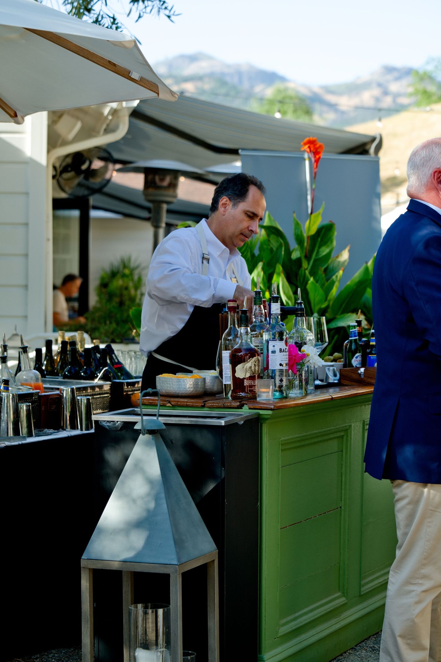 A bartender in white shirt and apron serves drinks at an outdoor bar setup with bottles and a green wood cabinet.