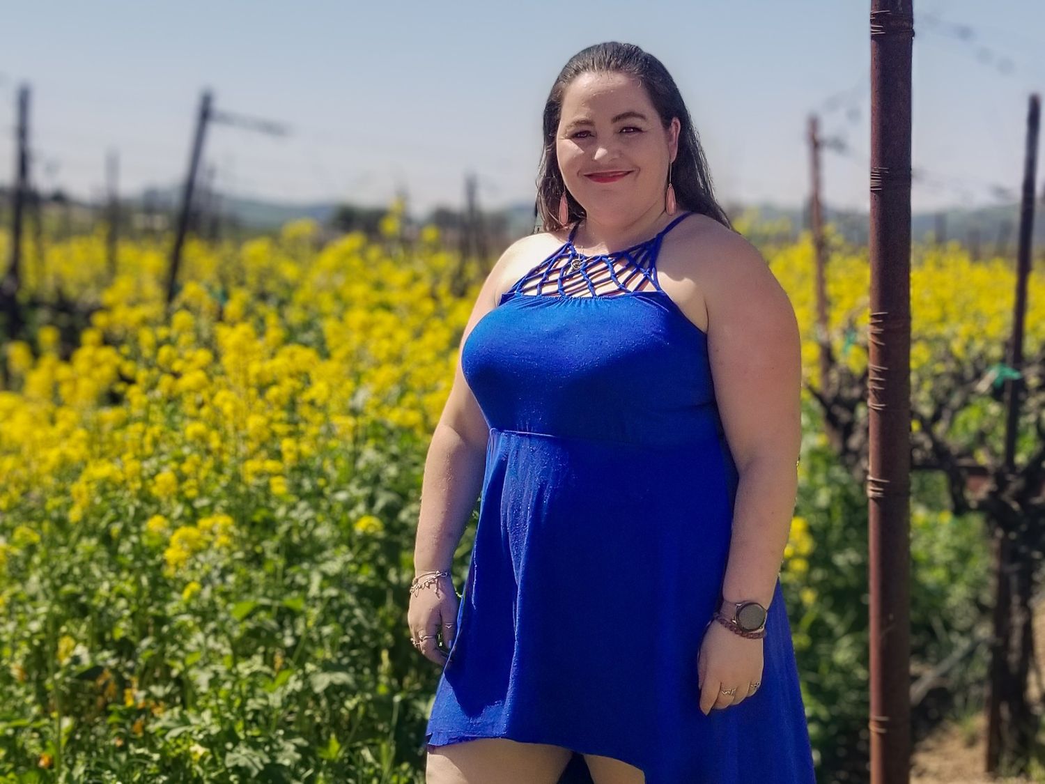 Woman in blue sundress poses in front of vibrant yellow wildflower field on sunny day.