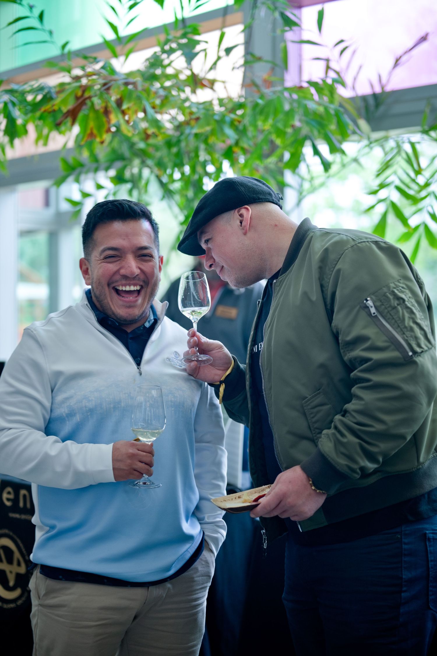 Two friends share a laugh while enjoying drinks together on an outdoor patio with green foliage in the background.