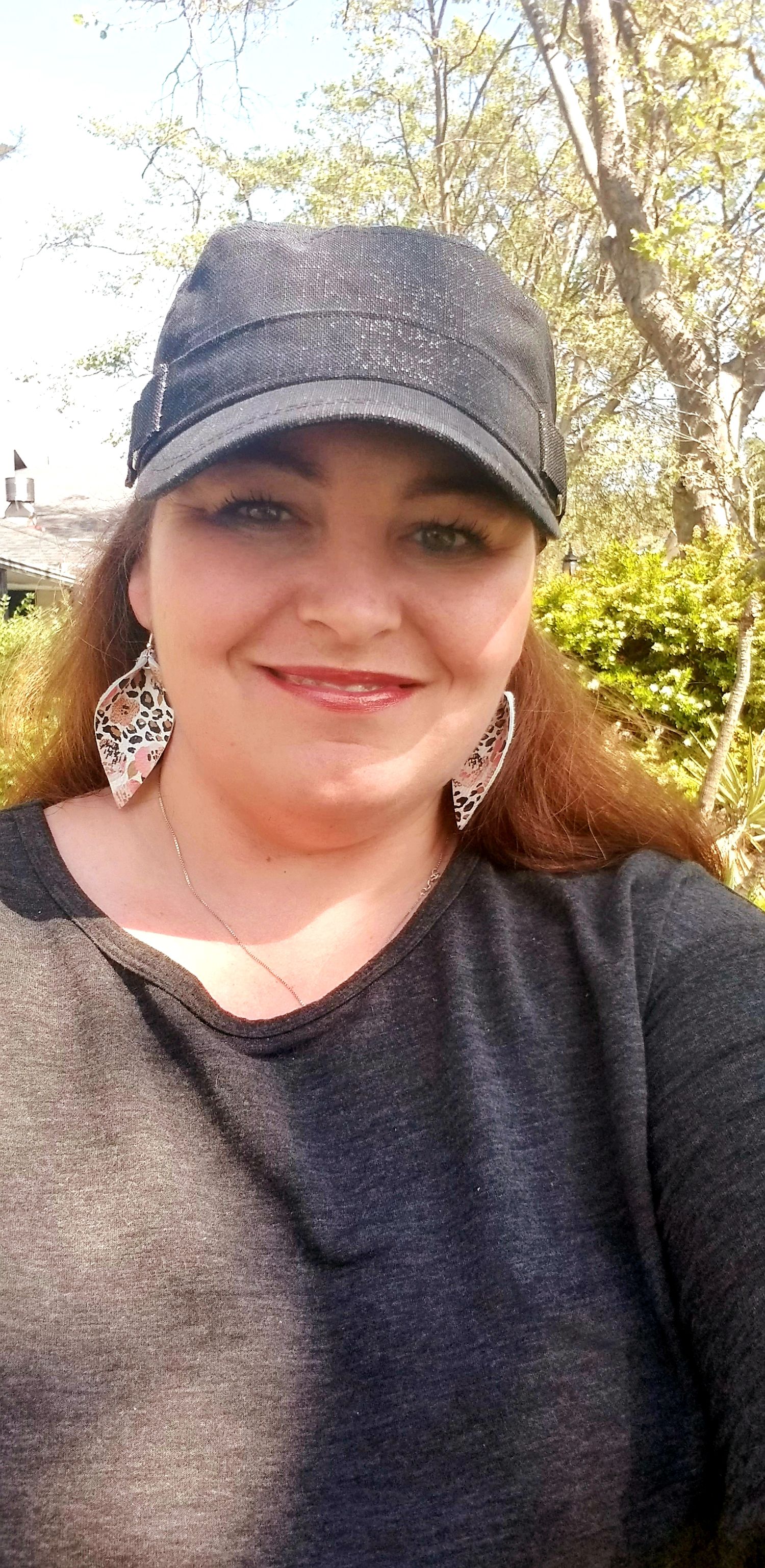 A person wearing a dark baseball cap and dark shirt smiles outdoors with trees visible in the background.