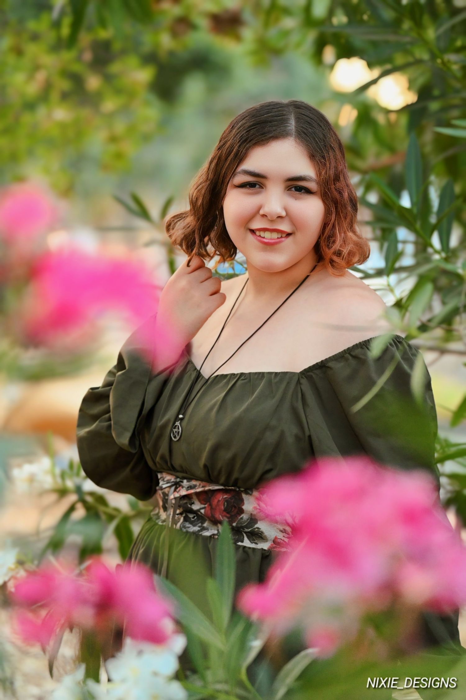 Senior girl in olive green dress poses outdoors among pink flowers and greenery against a soft bokeh background. Senior Portraits Napa, CA