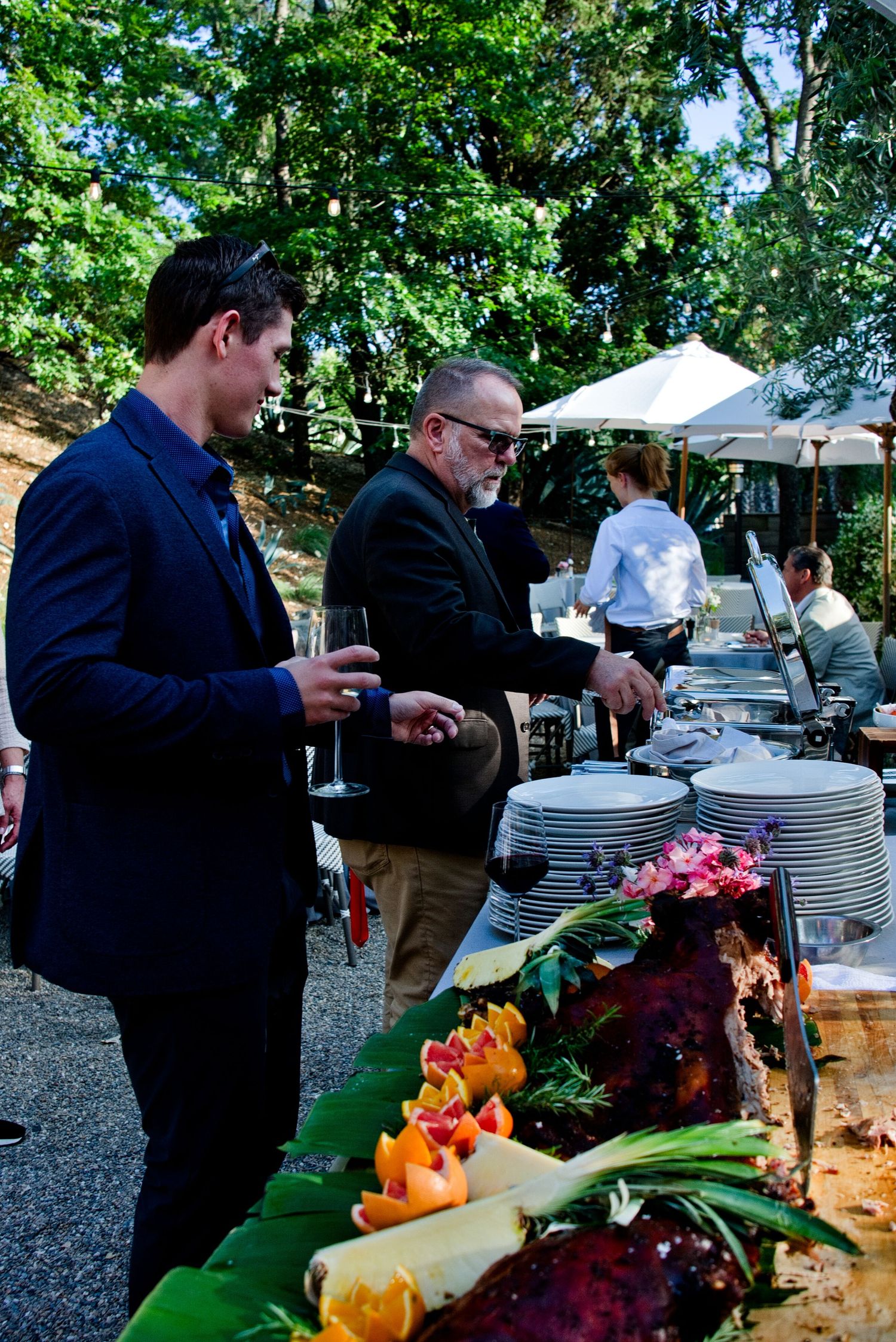Guests line up at an outdoor catering buffet with elegant table settings and colorful floral arrangements.