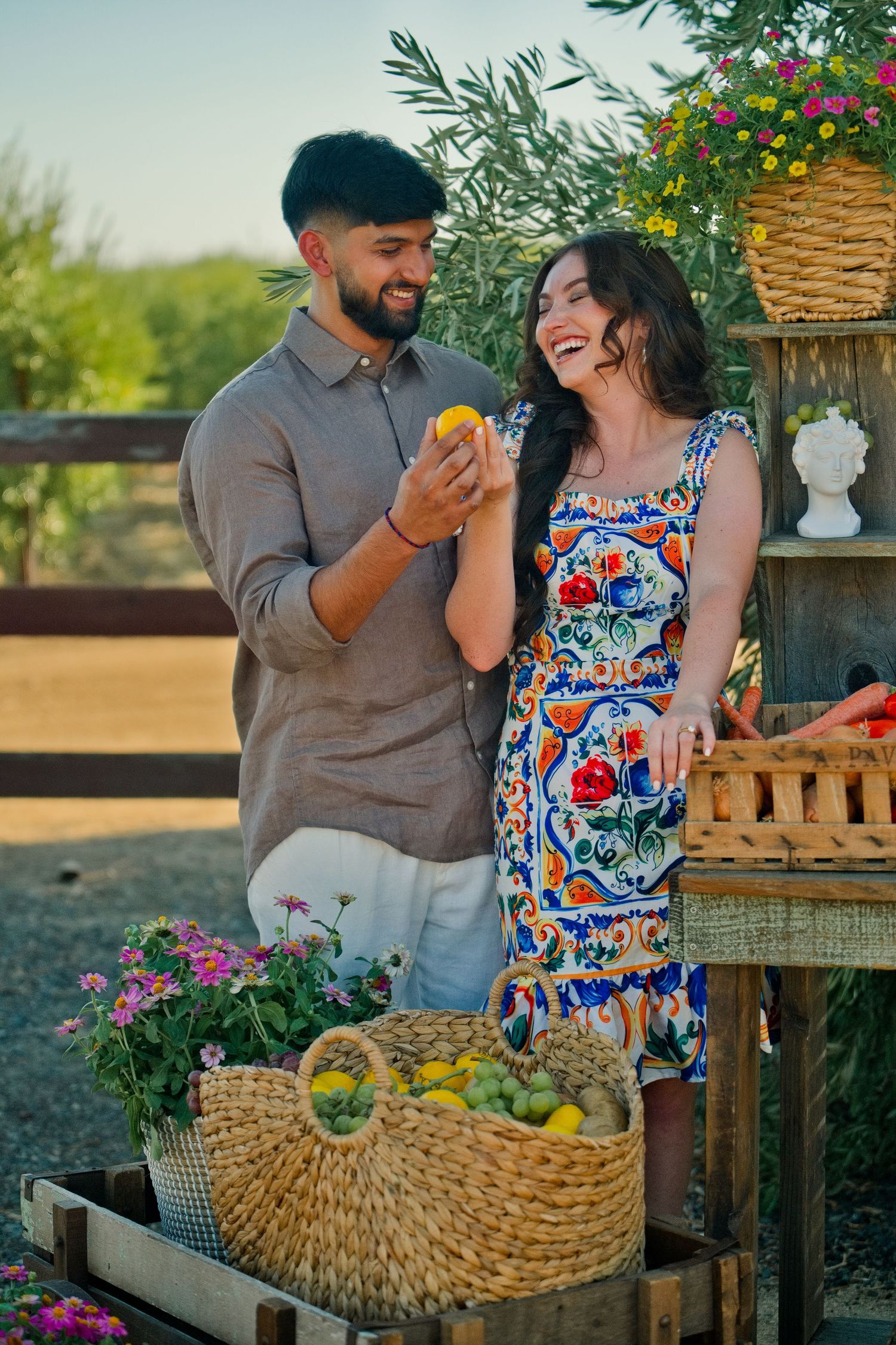 Happy couple sharing fresh produce at an outdoor market stand decorated with colorful flowers and woven baskets.