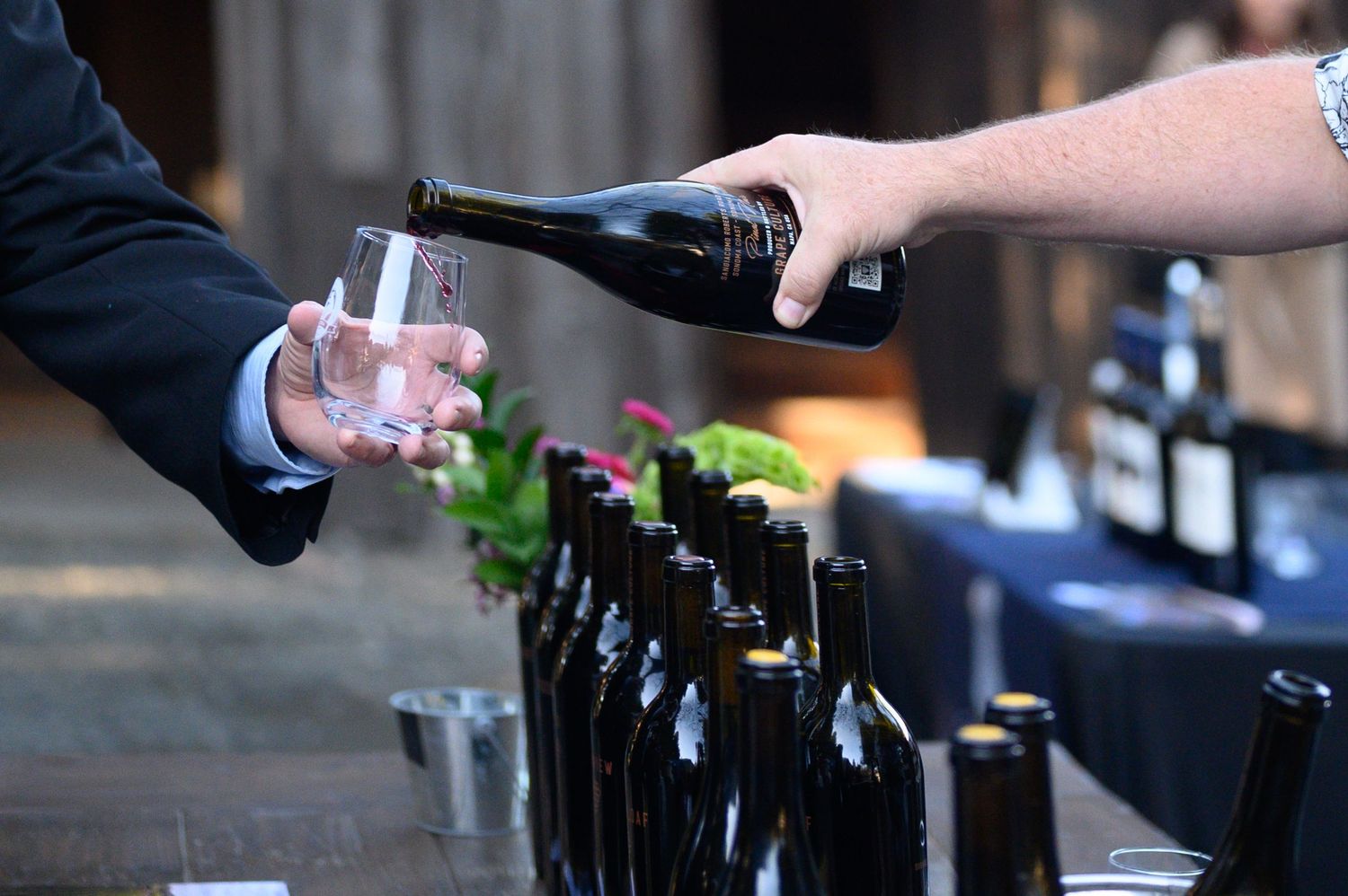 Wine being poured from a bottle into a glass at a tasting event with multiple bottles lined up on the bar.