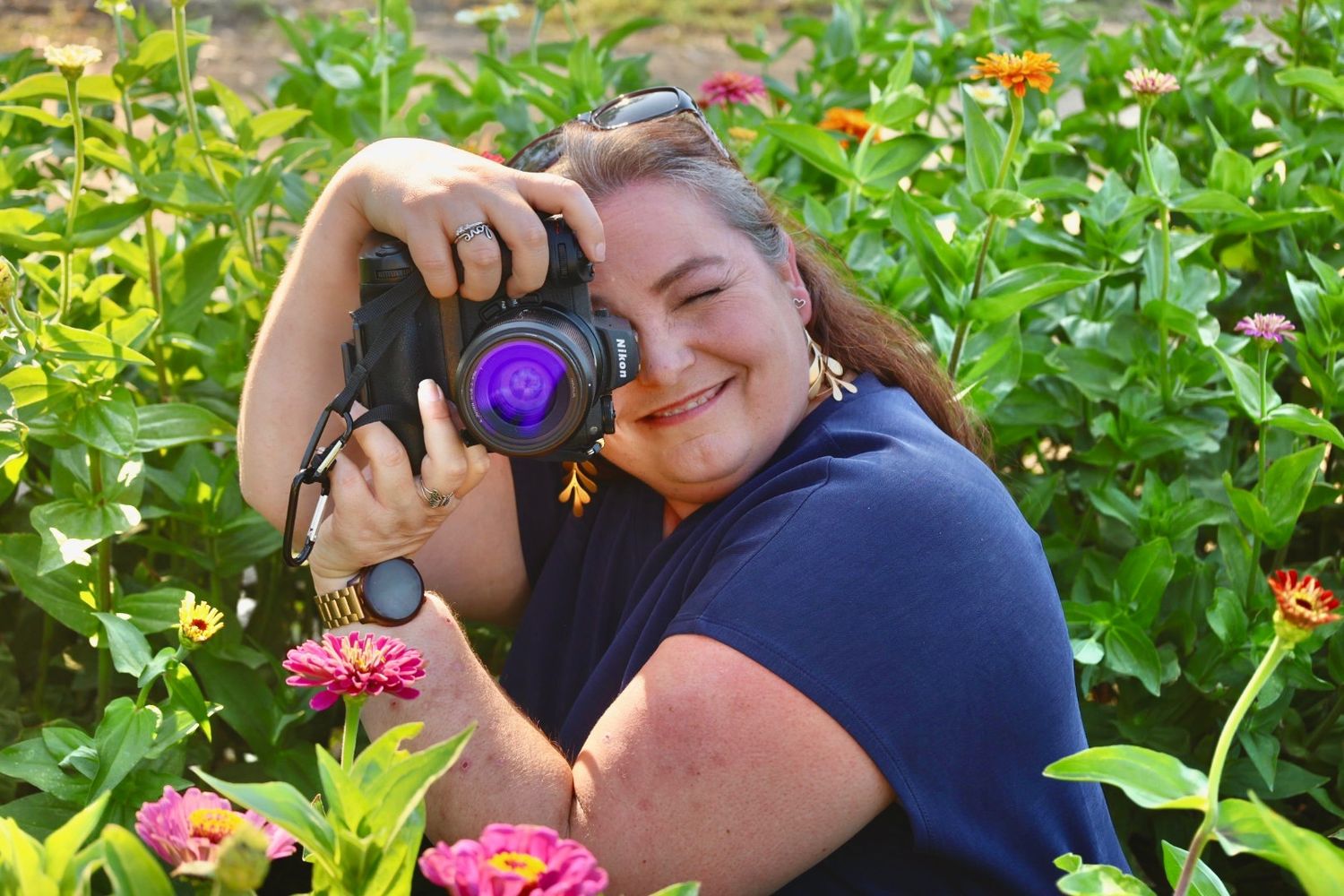 A photographer captures images in a garden filled with blooming zinnias and green foliage on a sunny day.
