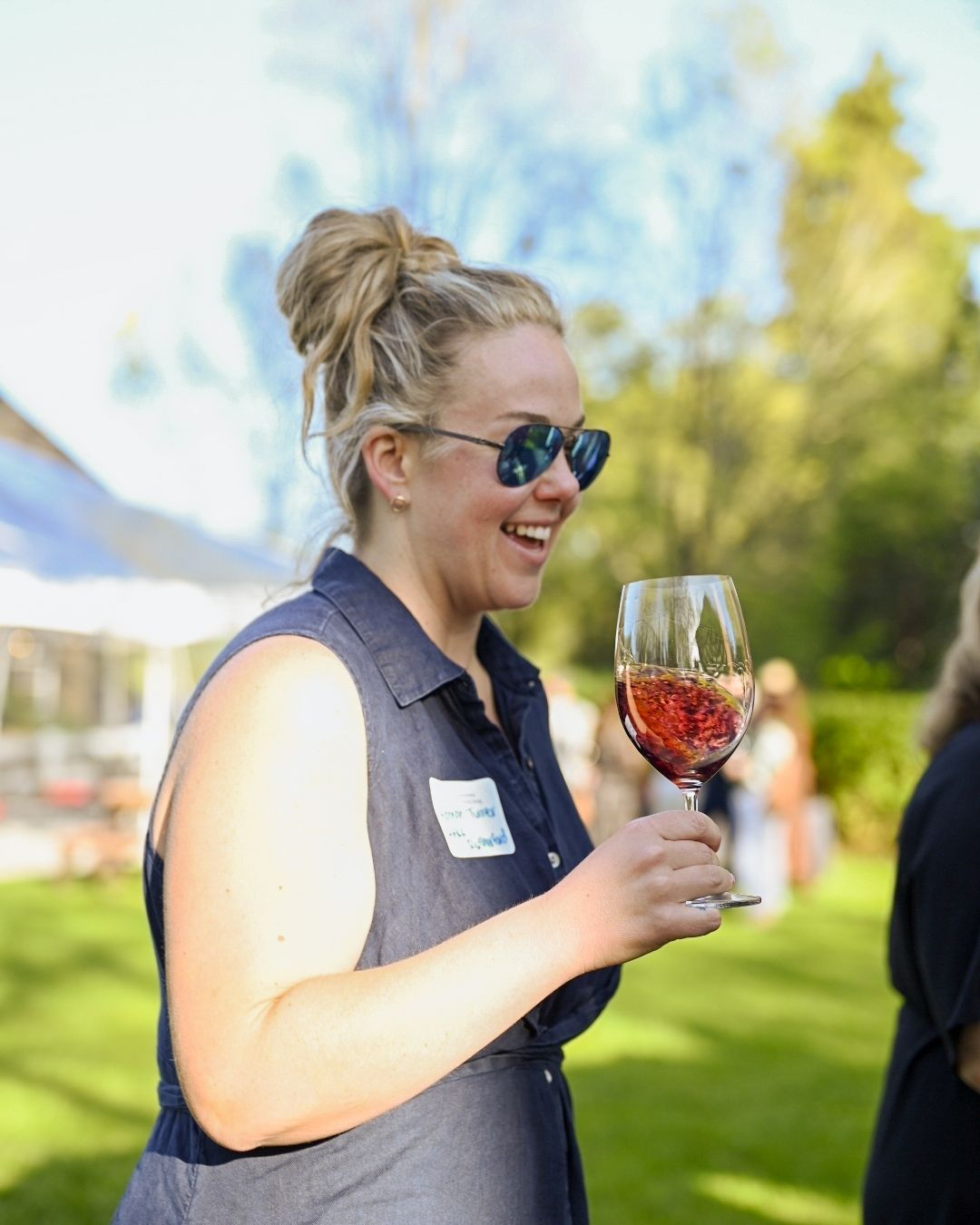 A person wearing sunglasses and a name tag enjoys a glass of red wine at an outdoor summer event.