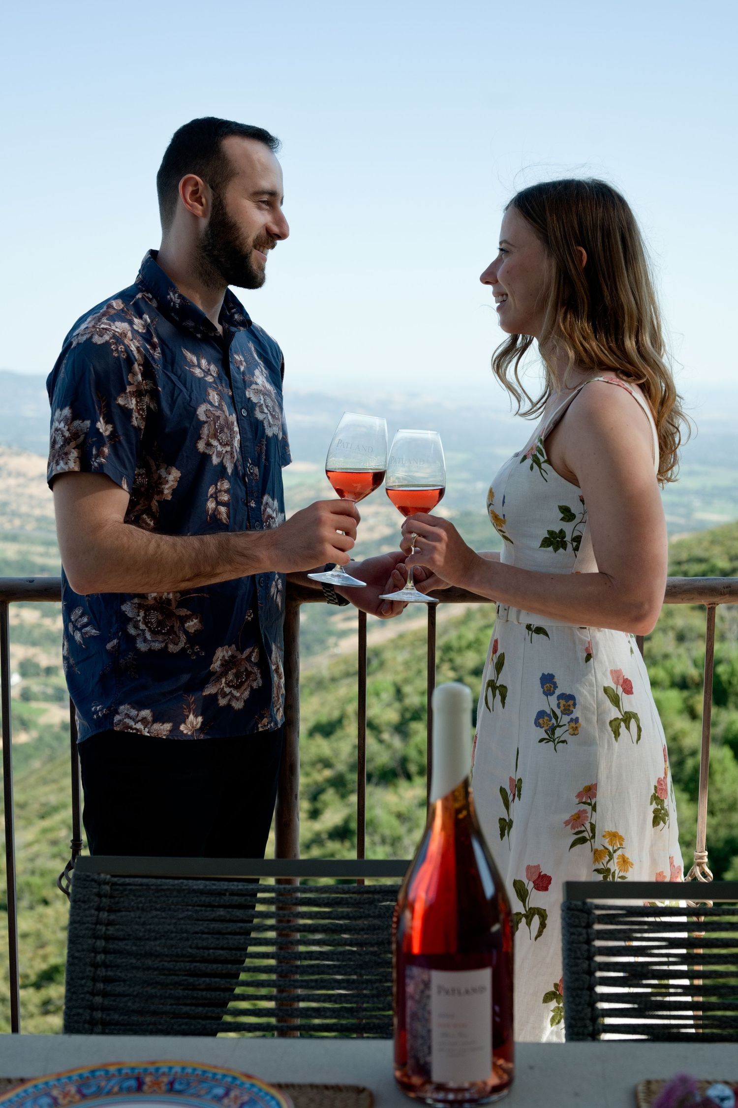 A couple shares wine and conversation on an outdoor balcony overlooking a scenic landscape at sunset.