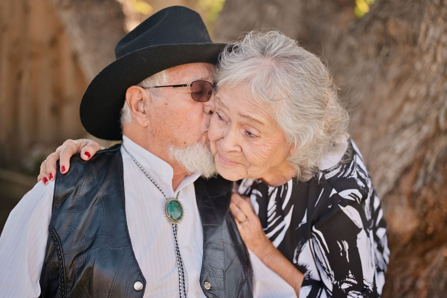 Elderly couple in western attire sharing an intimate moment outdoors with sunlight filtering through trees. Celebrating 72 years of marriage