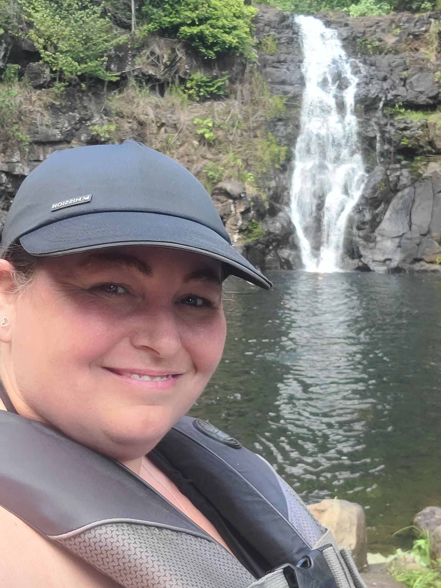Scenic waterfall cascades down rocky cliff into a dark pool surrounded by lush greenery and forest.