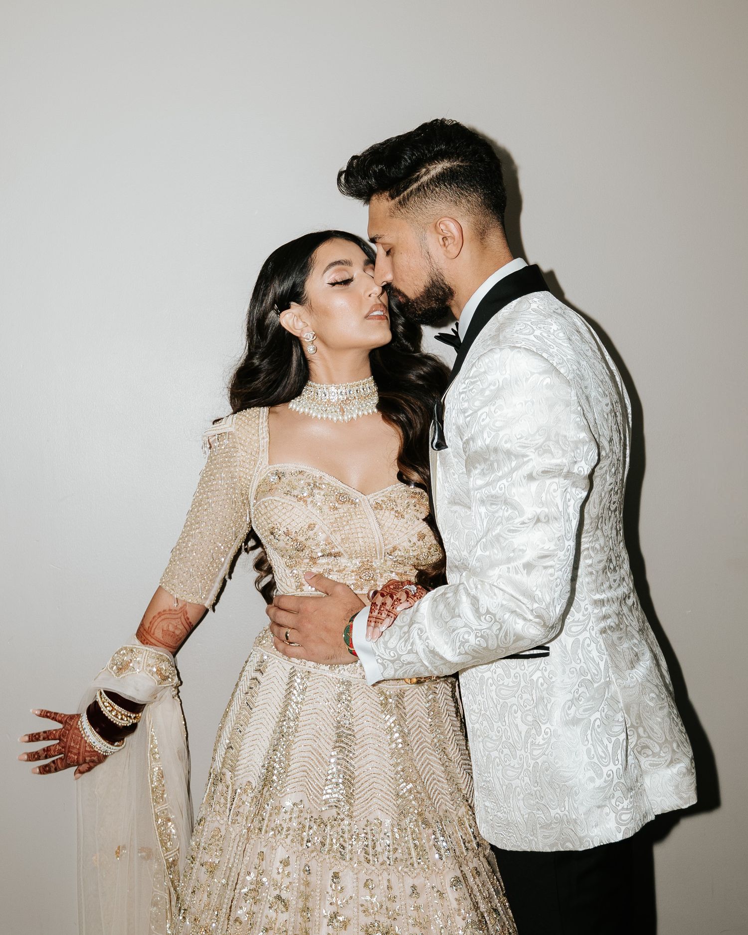 Couple in elegant wedding attire share intimate moment against white backdrop.