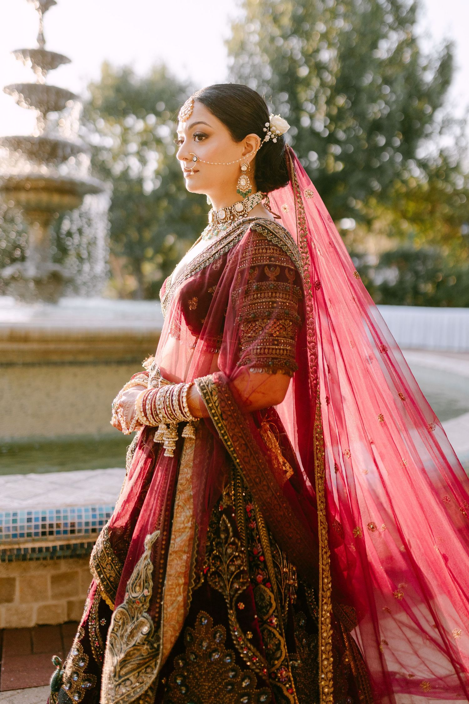 Bride in elaborate red and gold Indian lehenga poses elegantly by ornate fountain during outdoor wedding photoshoot.