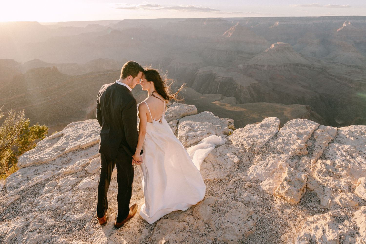 Bride and groom embrace on rocky Grand Canyon overlook at golden sunset, wedding dress flowing in wind.