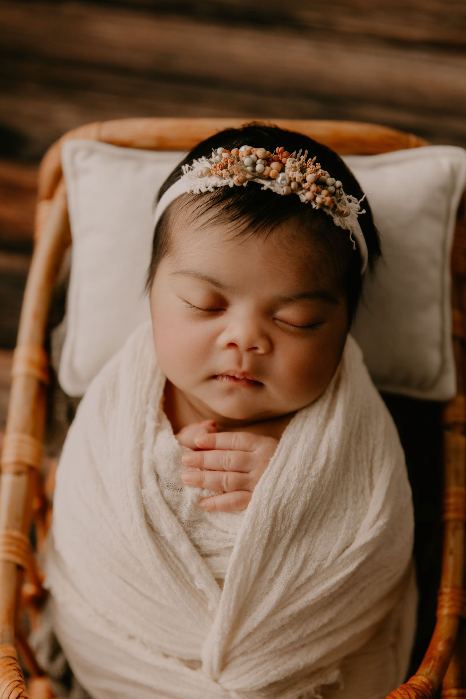 Sleeping newborn baby swaddled in white wrap wearing a delicate floral headband, resting peacefully in a wicker basket.