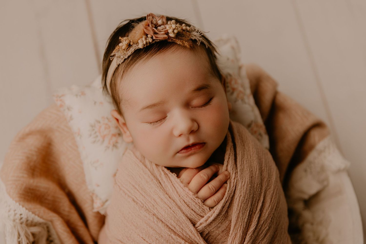 Sleeping newborn baby wrapped in soft peach swaddle with floral headband in cozy neutral-toned setting.