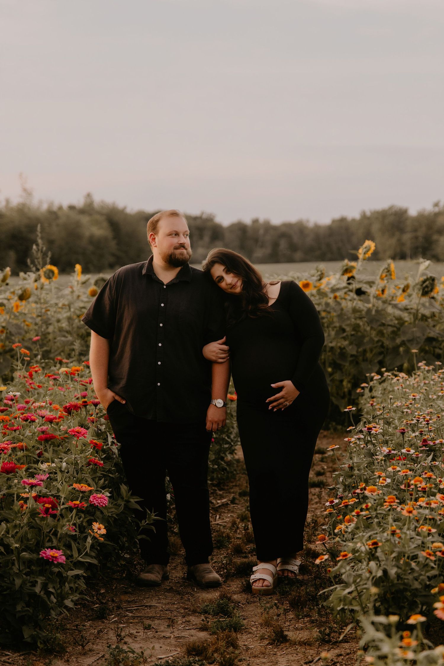 Couple in black outfits standing in a sunflower field at golden hour, woman leaning on man's shoulder.