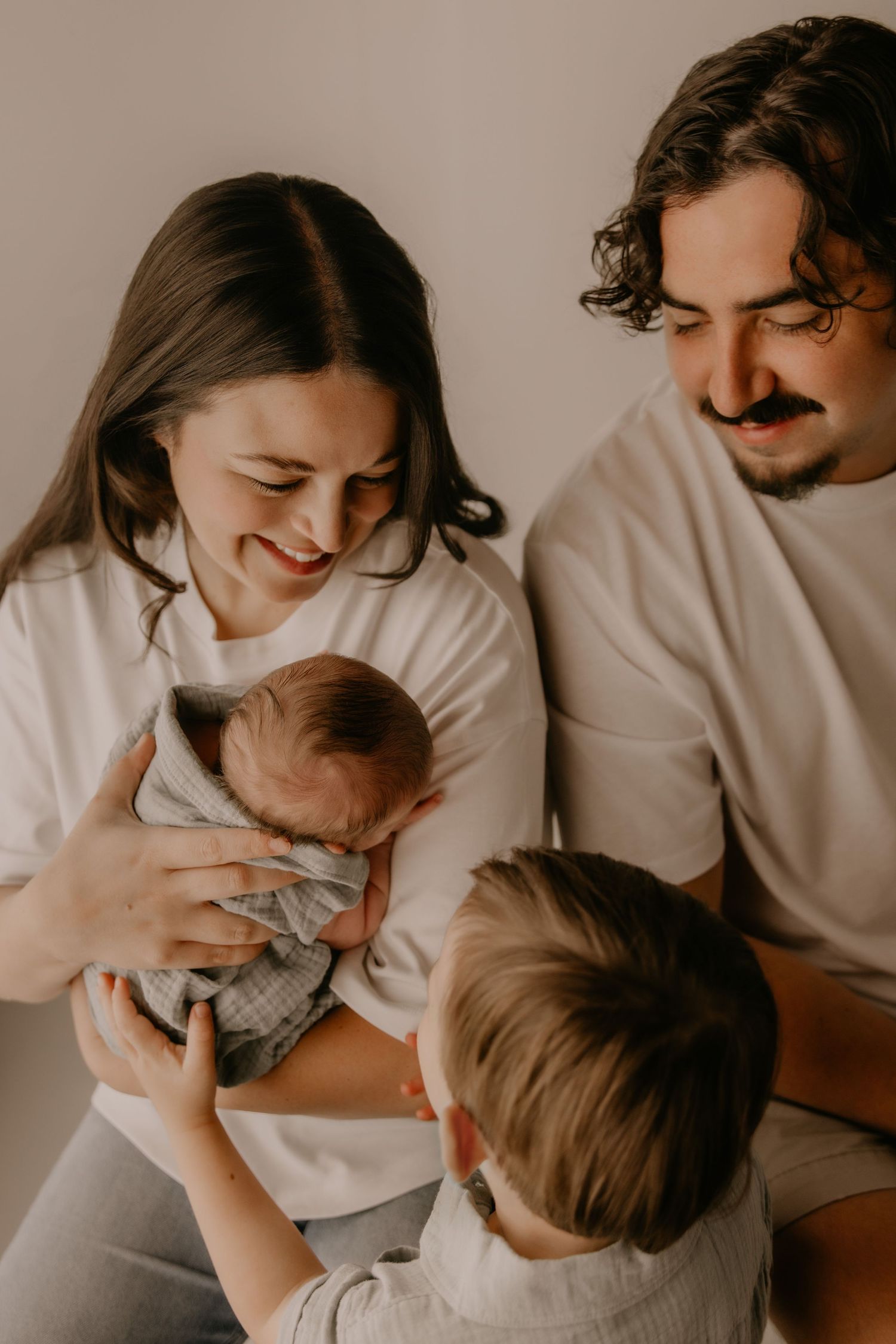 Happy family with parents smiling at newborn baby held by mother, with young child sitting nearby.