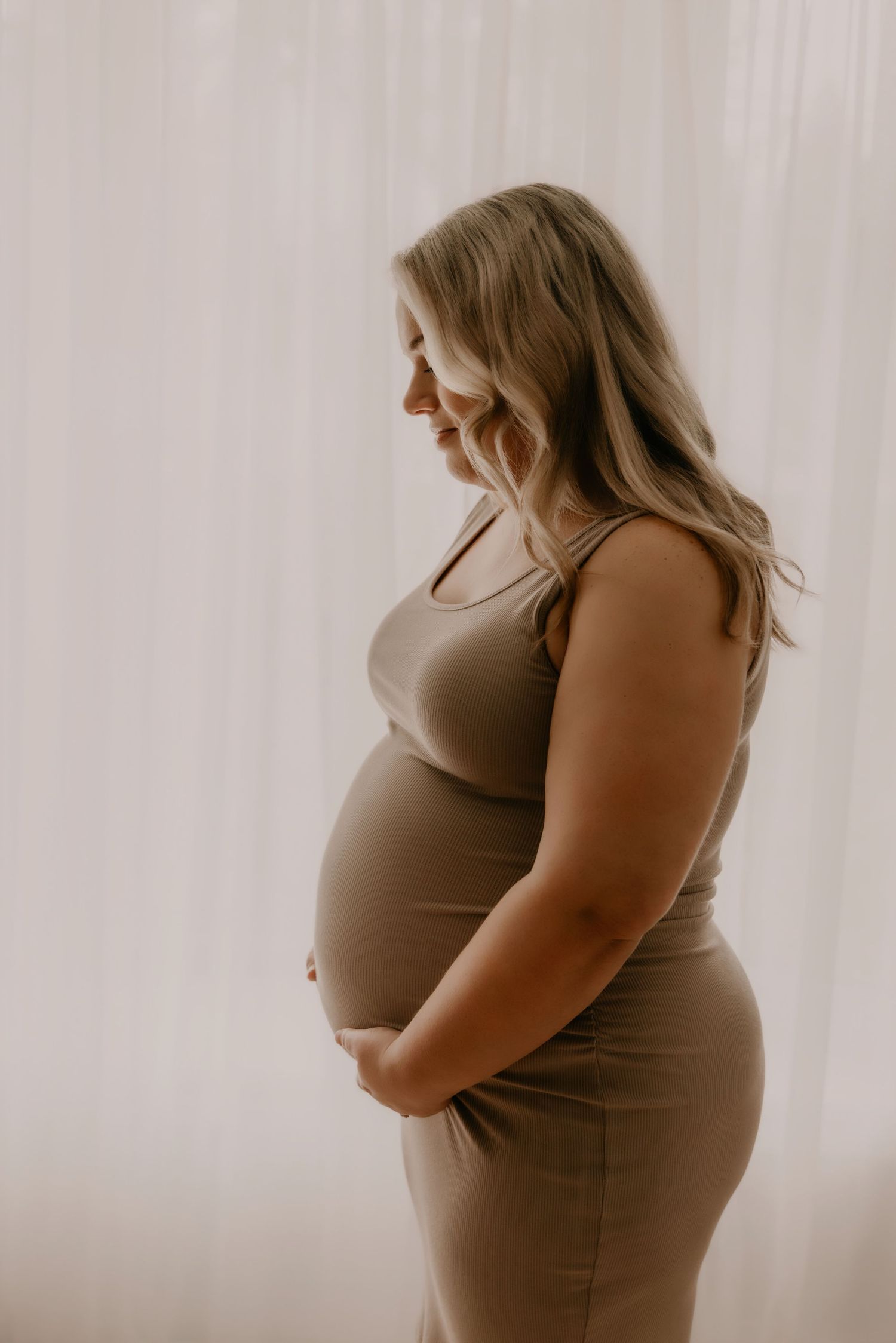 Pregnant woman in a tan dress cradling her baby bump, smiling softly in a serene, light-filled studio setting.