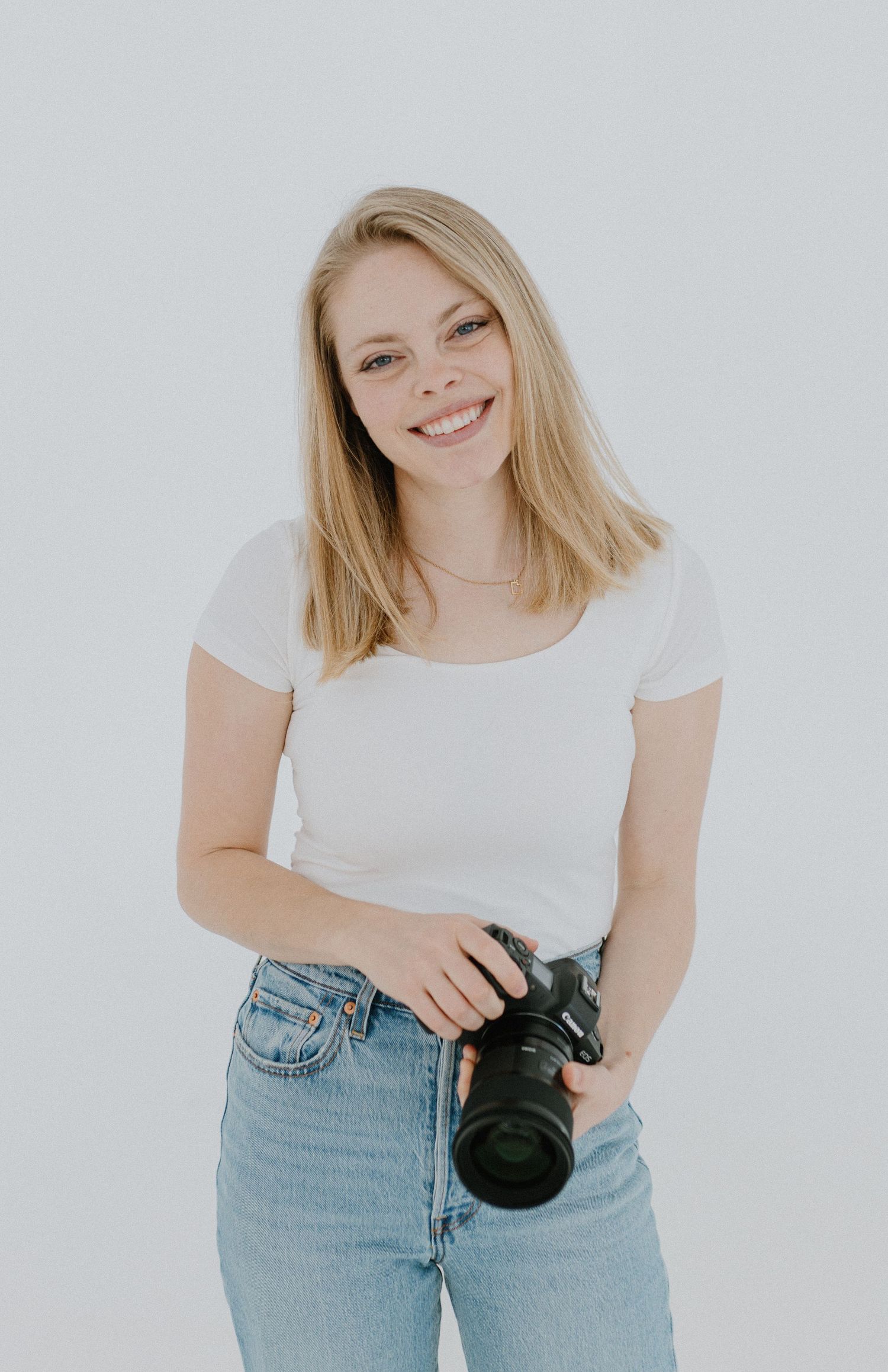 A photographer in casual attire with a white shirt and blue jeans holding a professional camera against a light background.