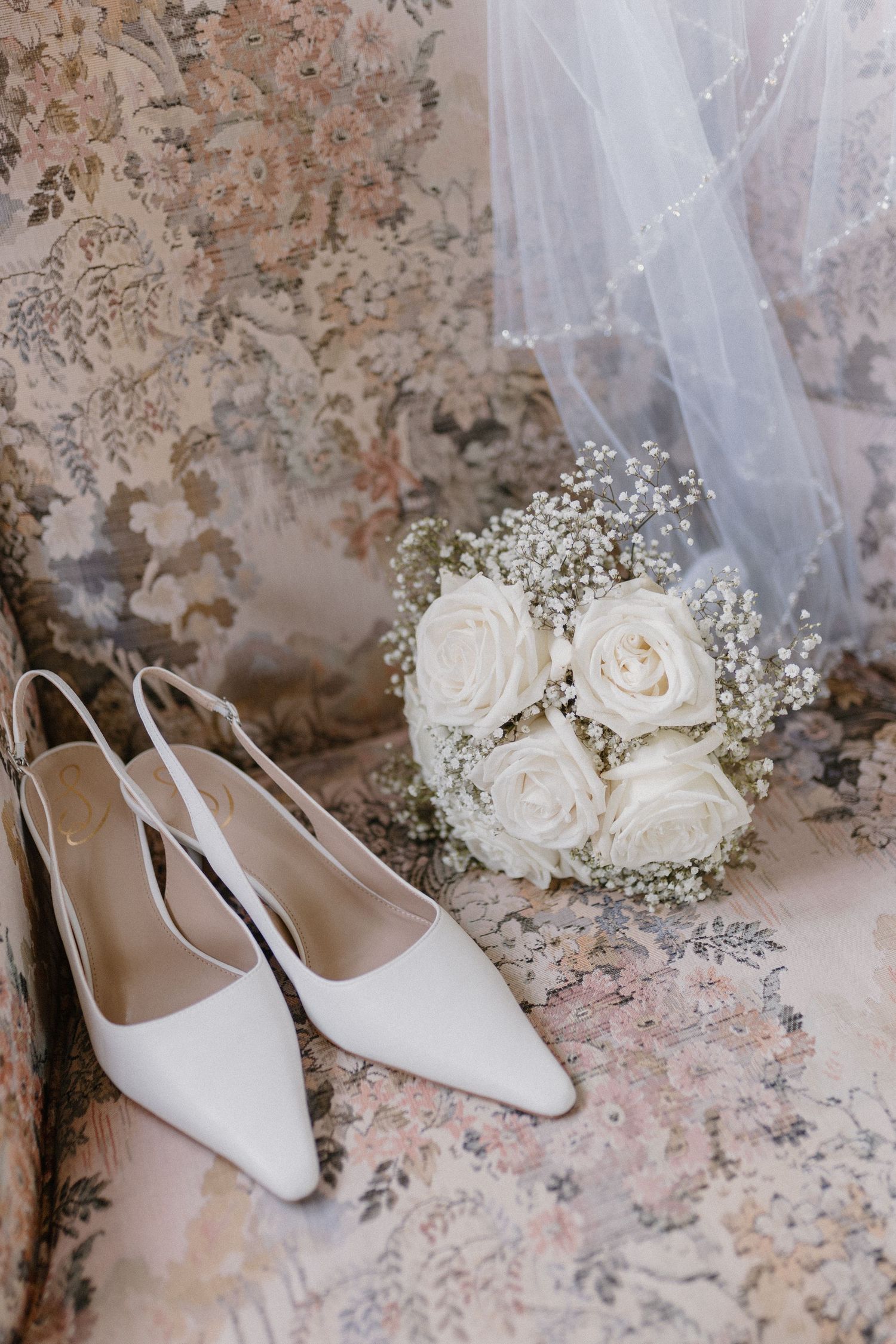 Elegant white bridal heels and delicate white rose bouquet with baby's breath against textured vintage wall.
