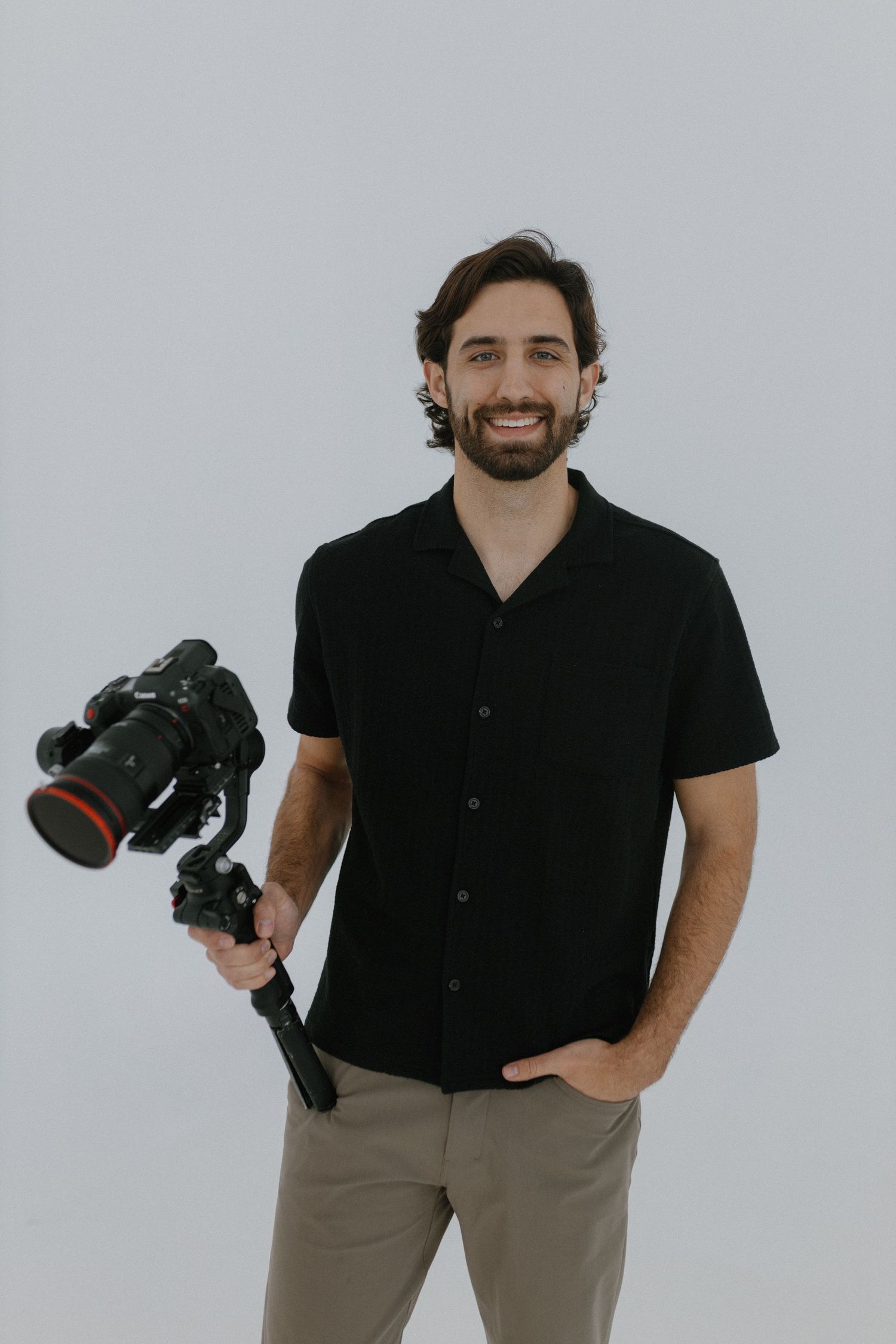 Professional videographer wearing black short-sleeve button-up shirt holds camera gimbal against white background.
