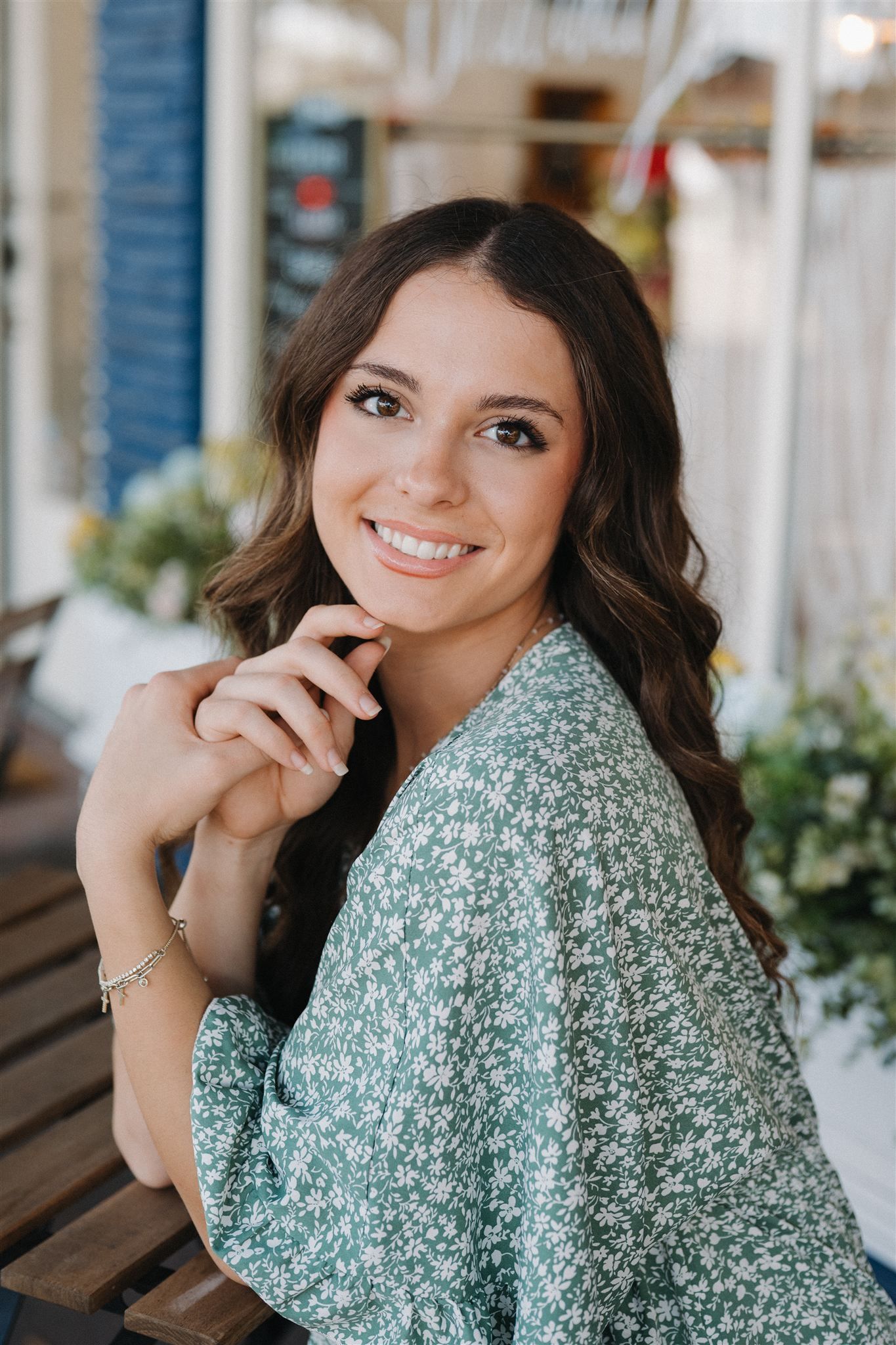 Portrait of senior girl in a green floral top sitting on a wooden bench outdoors with a natural smile.