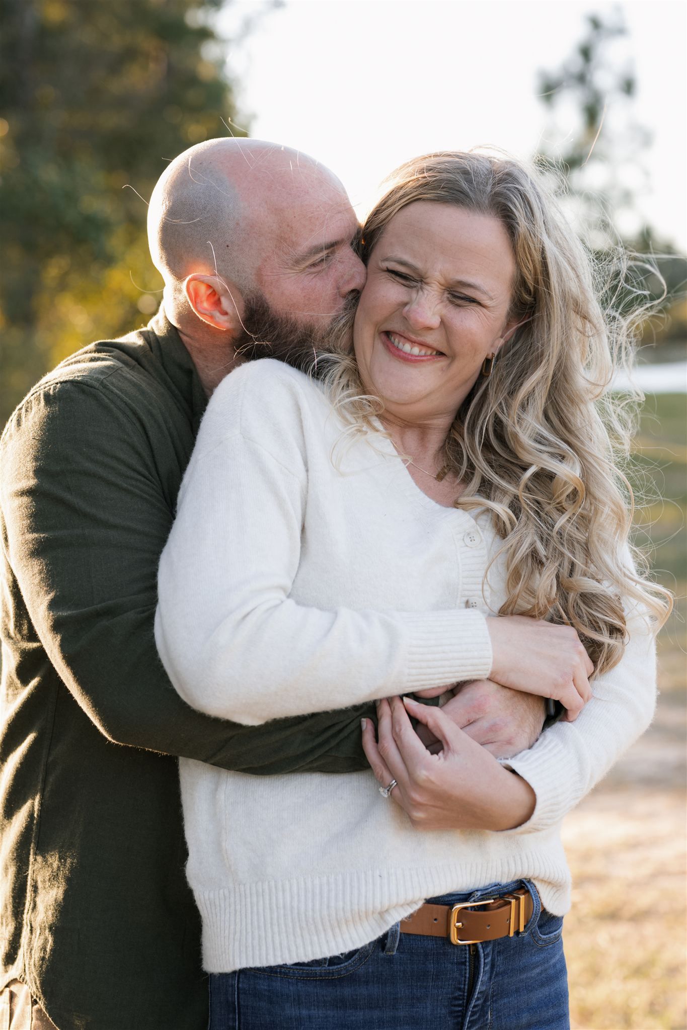 A bald bearded man hugs and kisses a smiling blonde woman in a white sweater outdoors in warm autumn light.