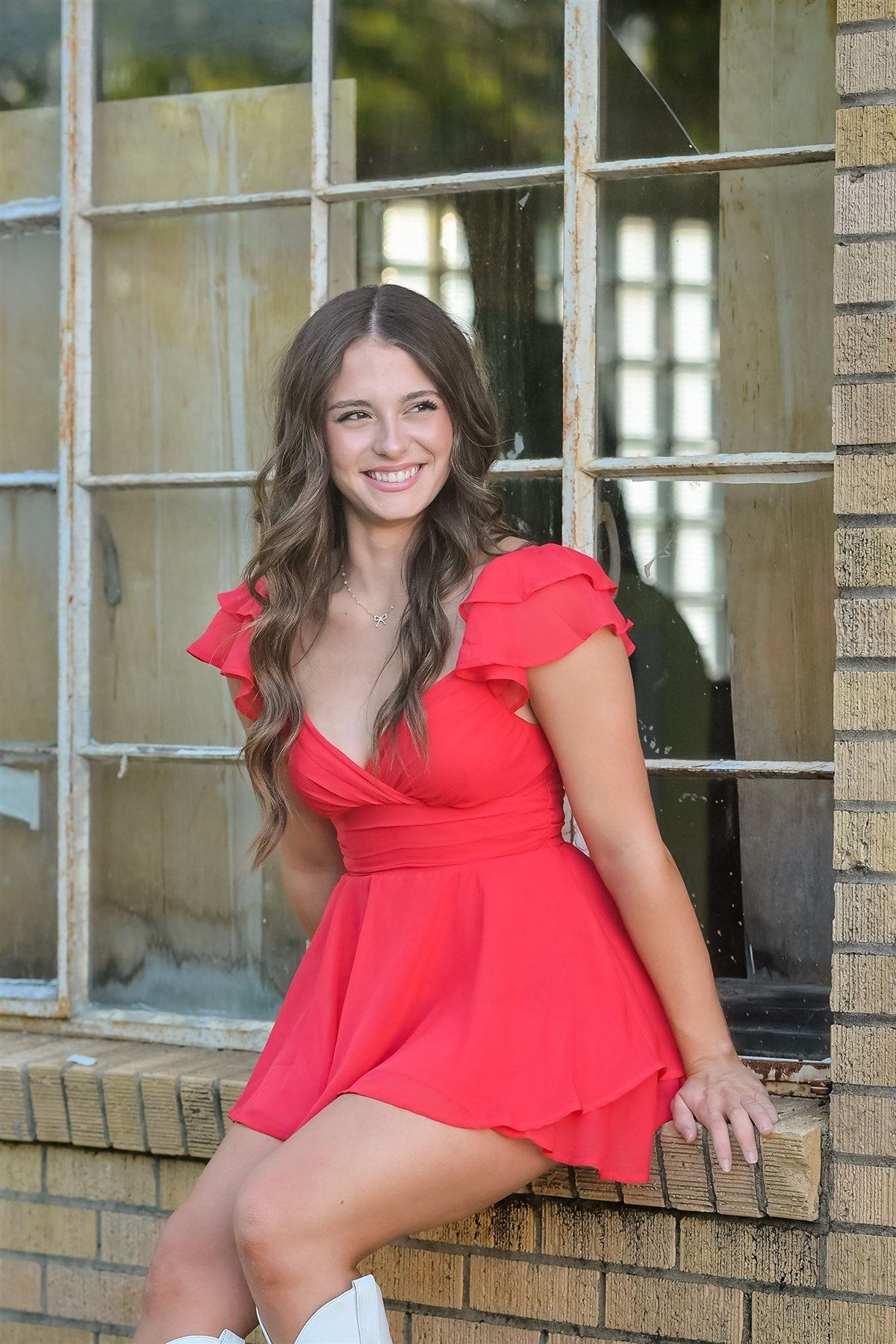 High school senior girl in bright red dress poses next to brick wall and window in Conroe, Tx.