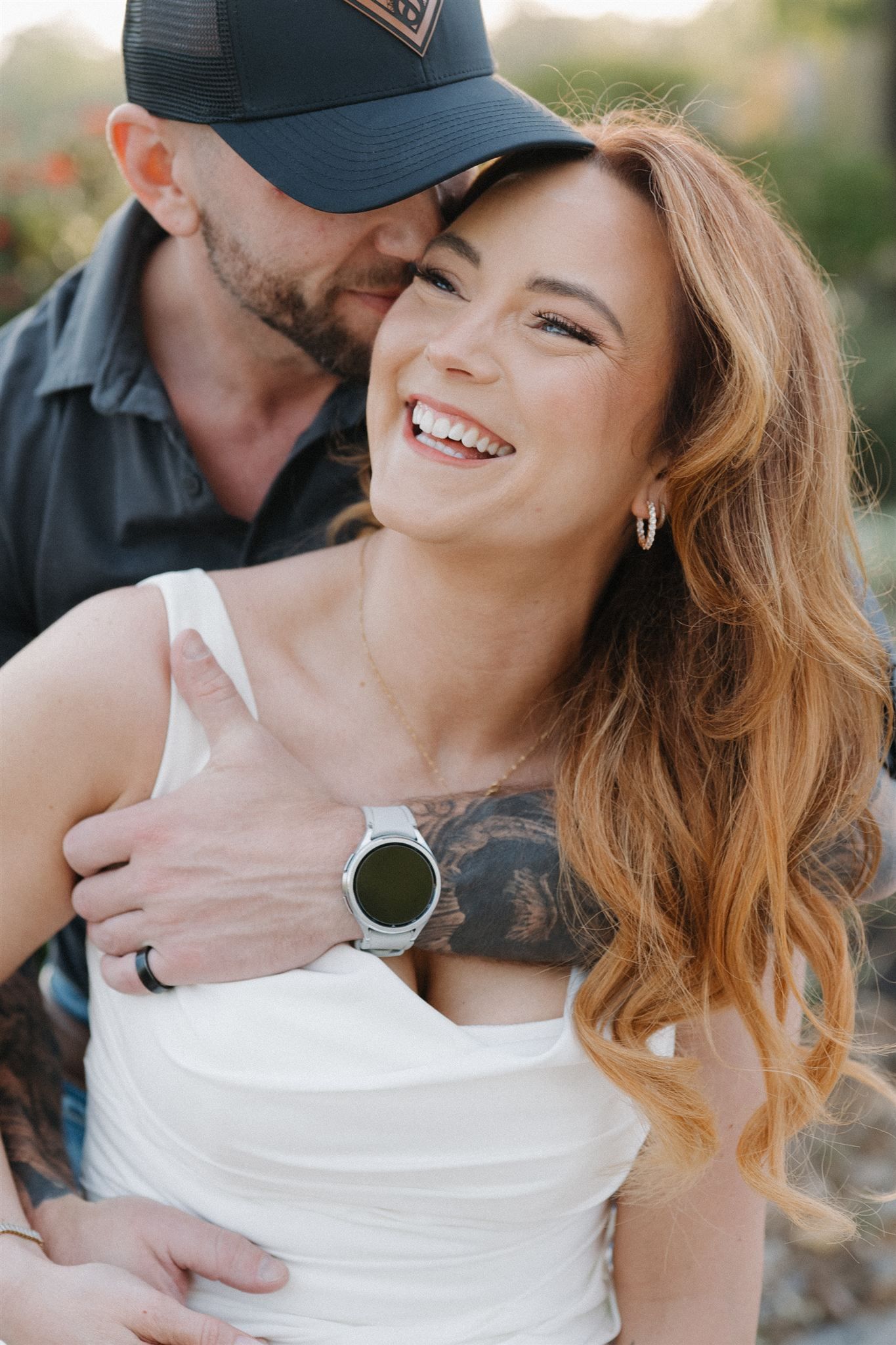 A smiling woman with auburn hair in a white top laughs as a man in a dark cap embraces her from behind outdoors.