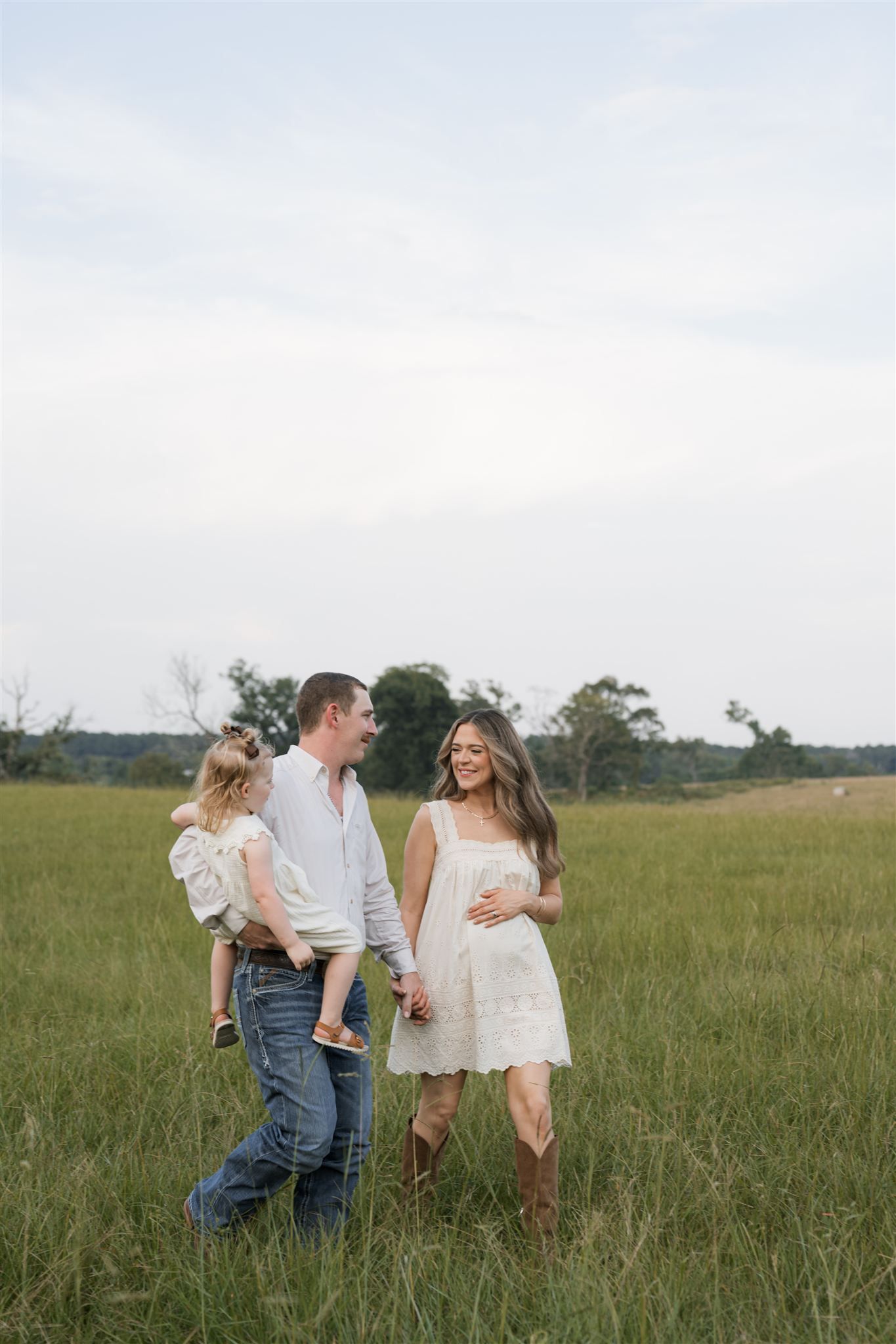 Parents and their young daughter walk and laugh together through a scenic summer meadow at sunset.