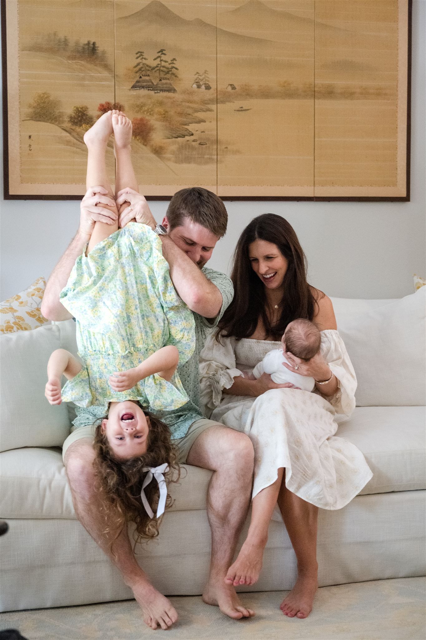 A playful family moment on a white couch with a mother holding their newborn and dad holding the toddler upside down.