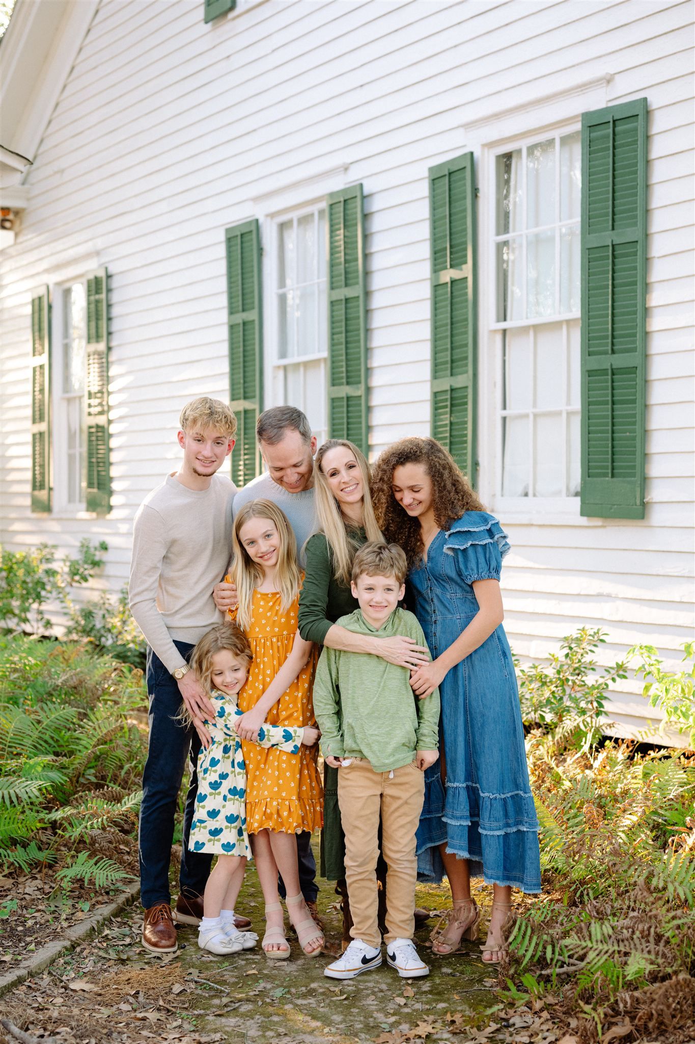 A group stands together outside a white farmhouse with green shutters and a garden of wildflowers.