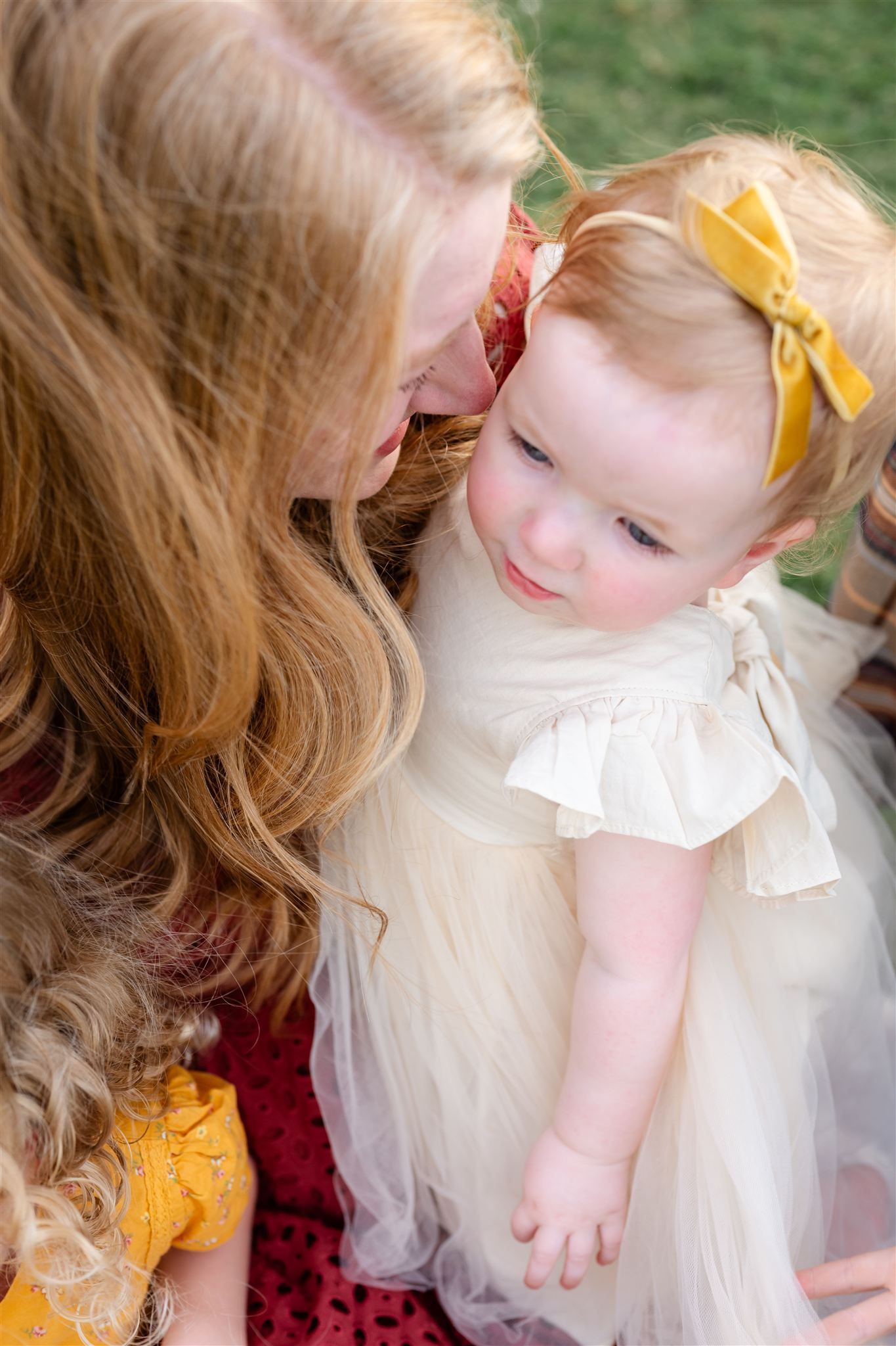 A tender moment shared between a woman with wavy hair and a baby wearing a yellow bow and white tulle dress.