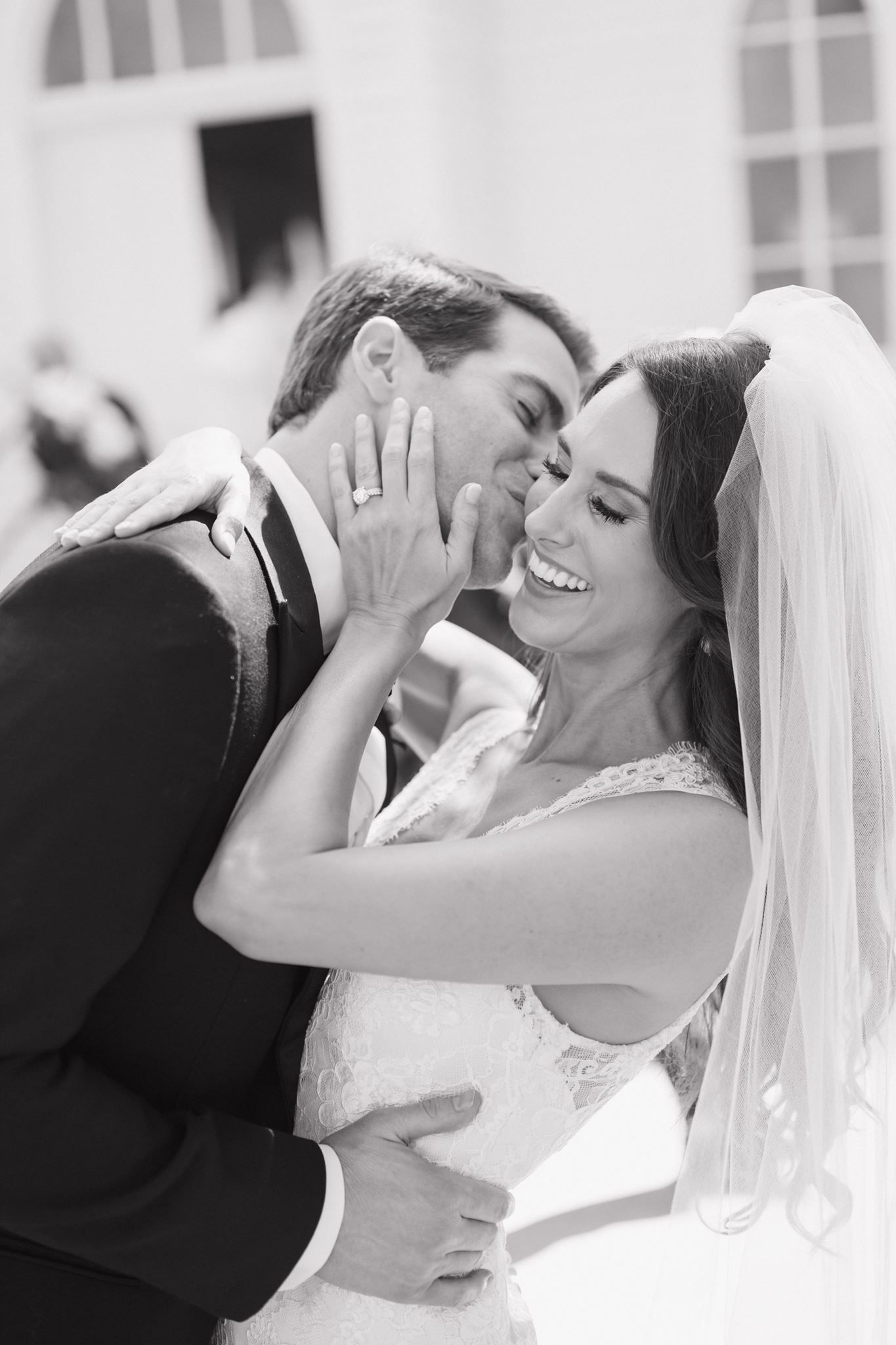 Black and white photo of a joyful bride laughing as groom kisses her cheek, showcasing her wedding ring and veil.