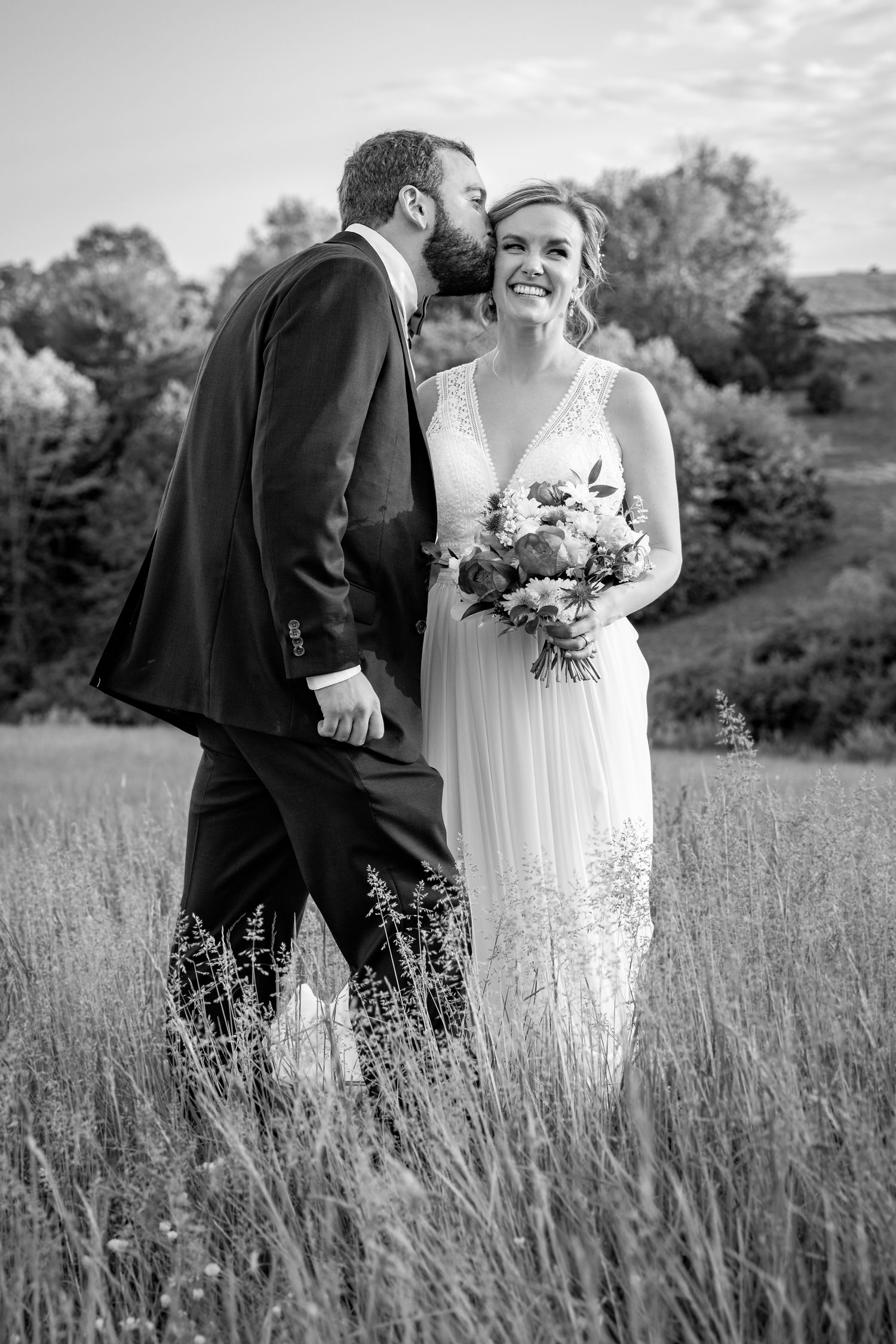 Bride and groom in a field with groom kissing bride on the cheek after their wedding ceremony..