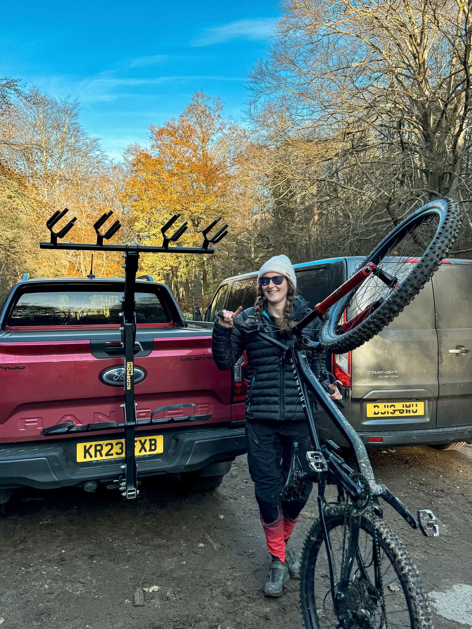 Mountain biker lifts bicycle next to red pickup truck with bike rack in autumn forest setting.