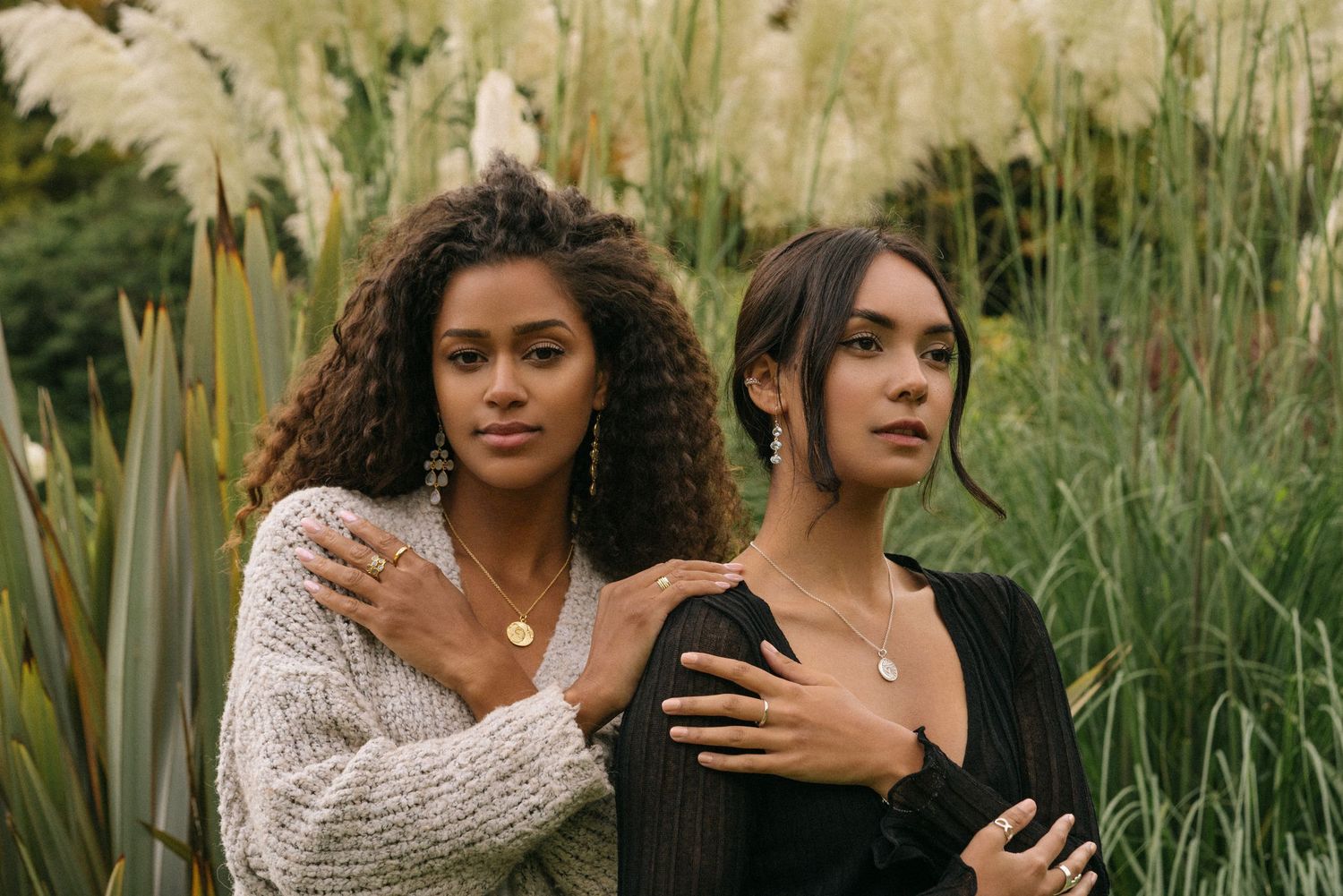 Two friends in knitwear pose together against tall ornamental grasses in an outdoor natural setting.