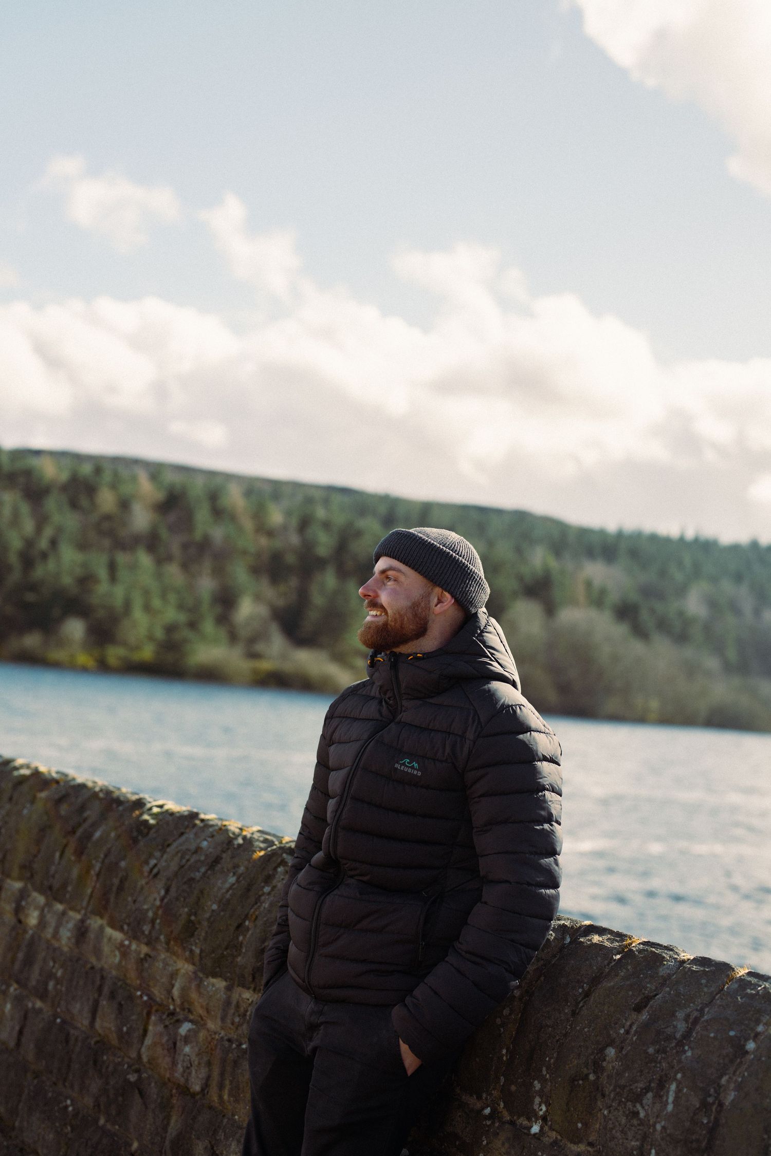 Someone wearing a black puffer jacket and beanie stands by a stone wall overlooking a scenic lake and forested hills.