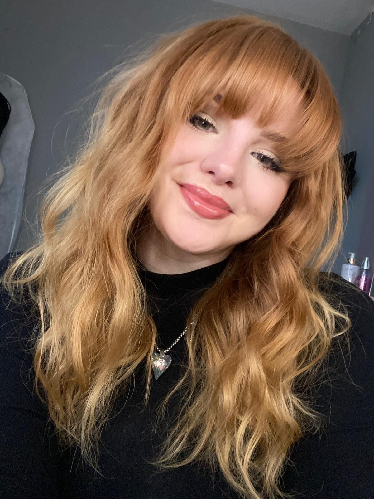 A selfie showing wavy strawberry blonde hair, red lipstick, and a black sweater with a necklace.