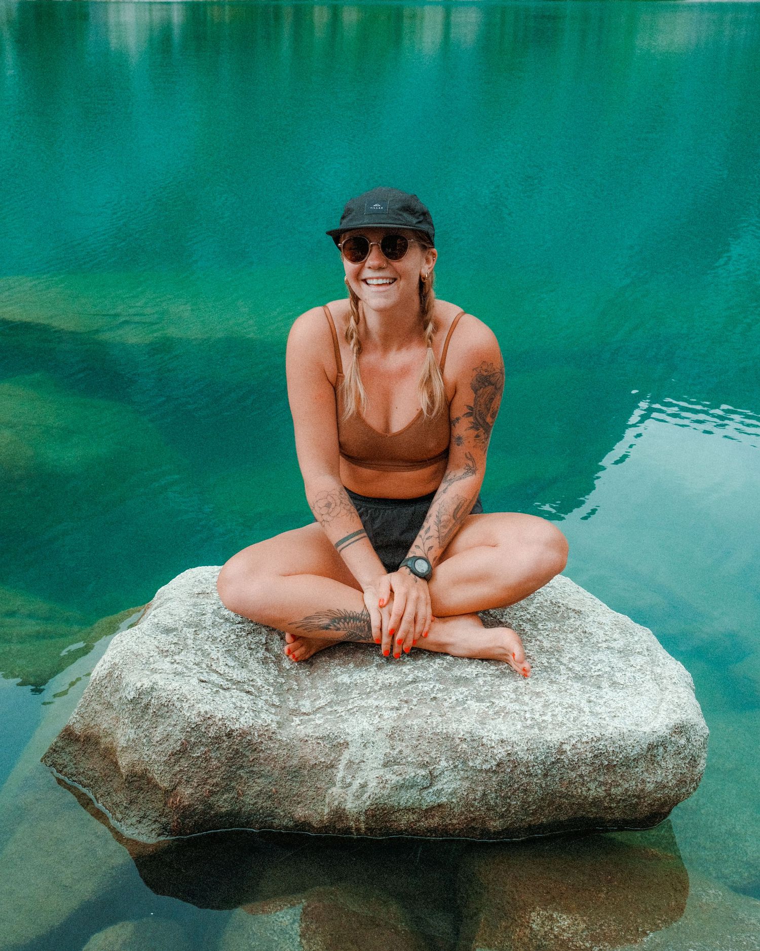 Woman in brown swimsuit sitting cross-legged on boulder by turquoise lake wearing cap and sunglasses.