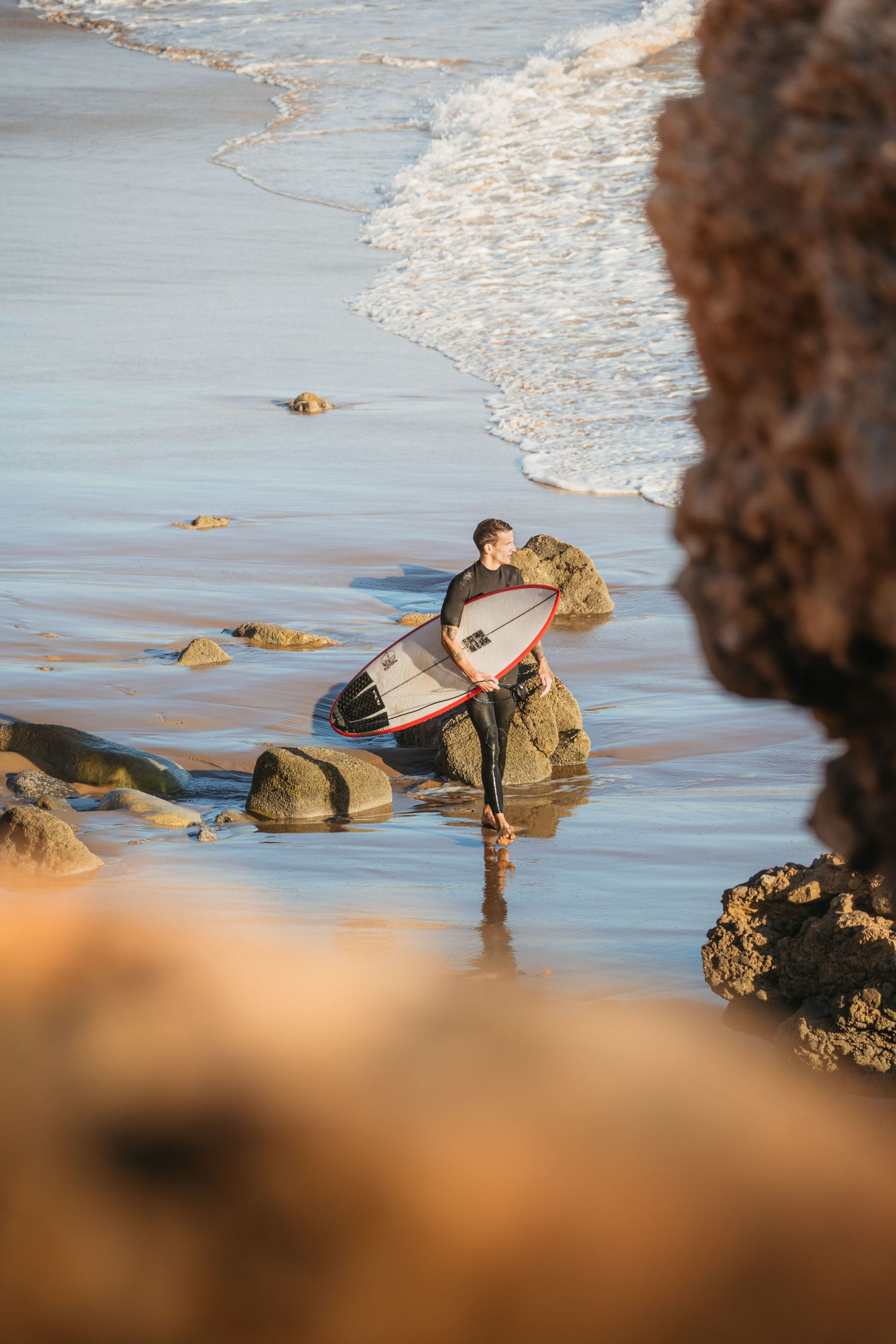 A surfer in a wetsuit walks along a rocky beach with a surfboard during golden hour.