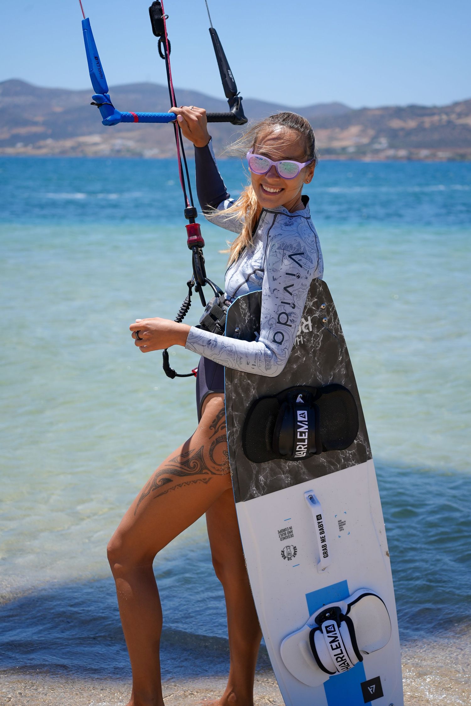 Kiteboarder in water sports gear smiles while holding equipment on a sunny beach with blue ocean and mountains in background.