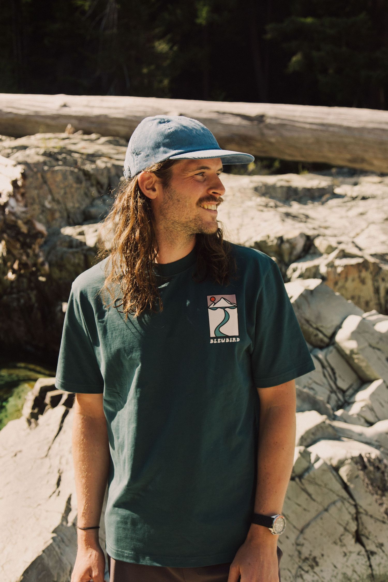 Person wearing a dark green t-shirt and baseball cap sits against a rocky outdoor backdrop in natural lighting.