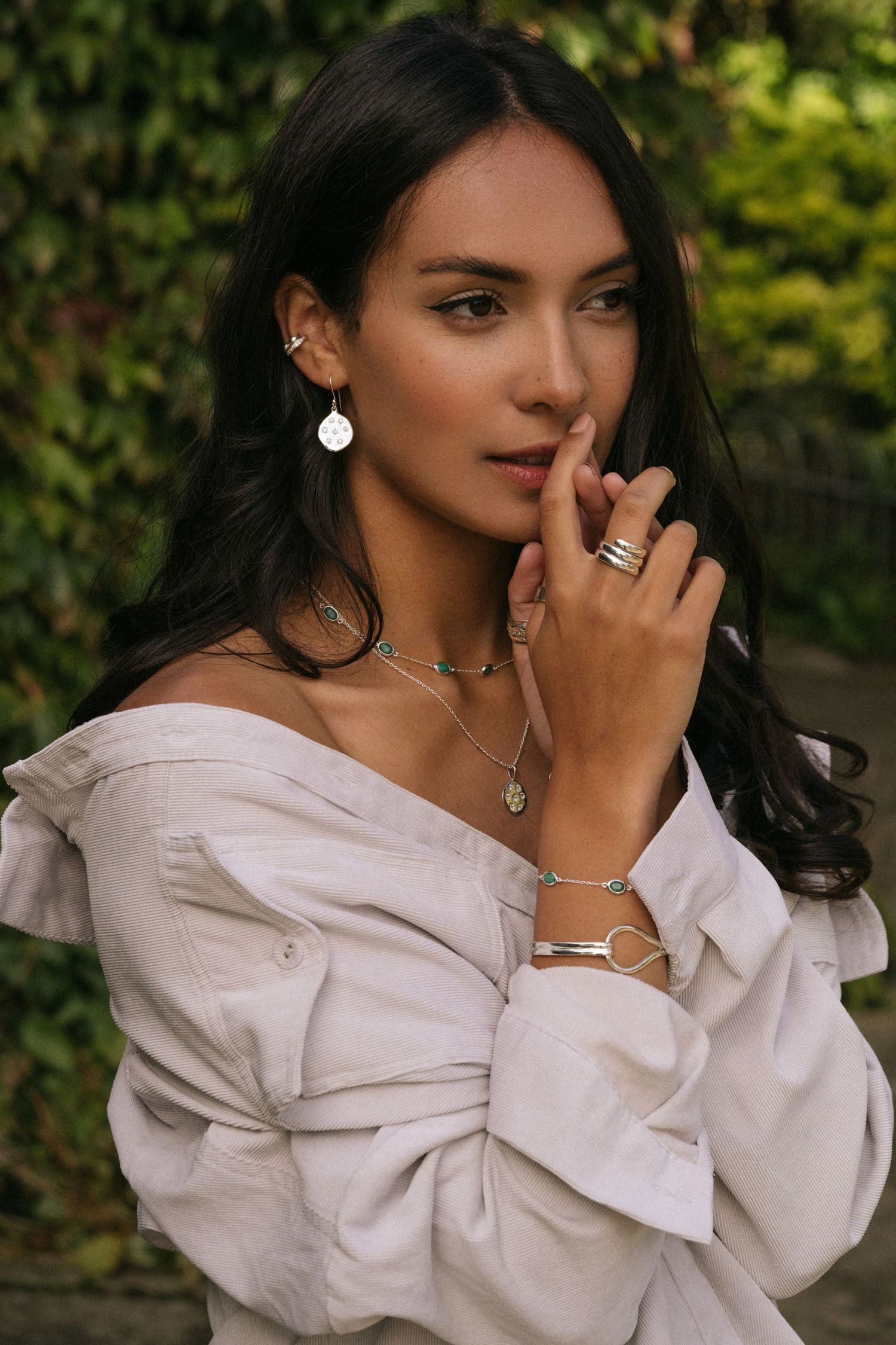 Person wearing white off-shoulder top with silver jewelry and dark hair poses outdoors against greenery.