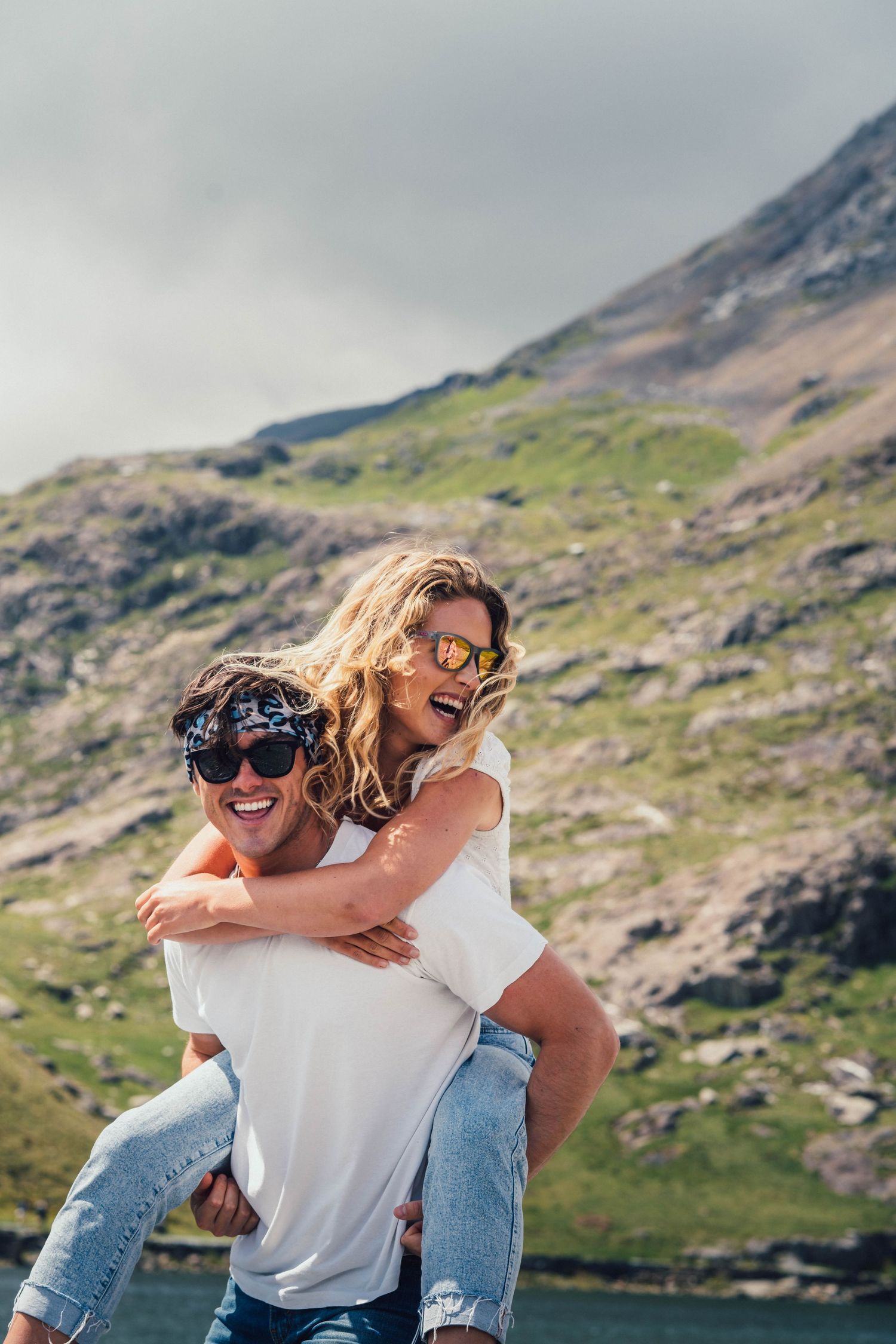Two friends enjoy a carefree piggyback moment against a scenic mountain landscape with green slopes and rocky terrain.
