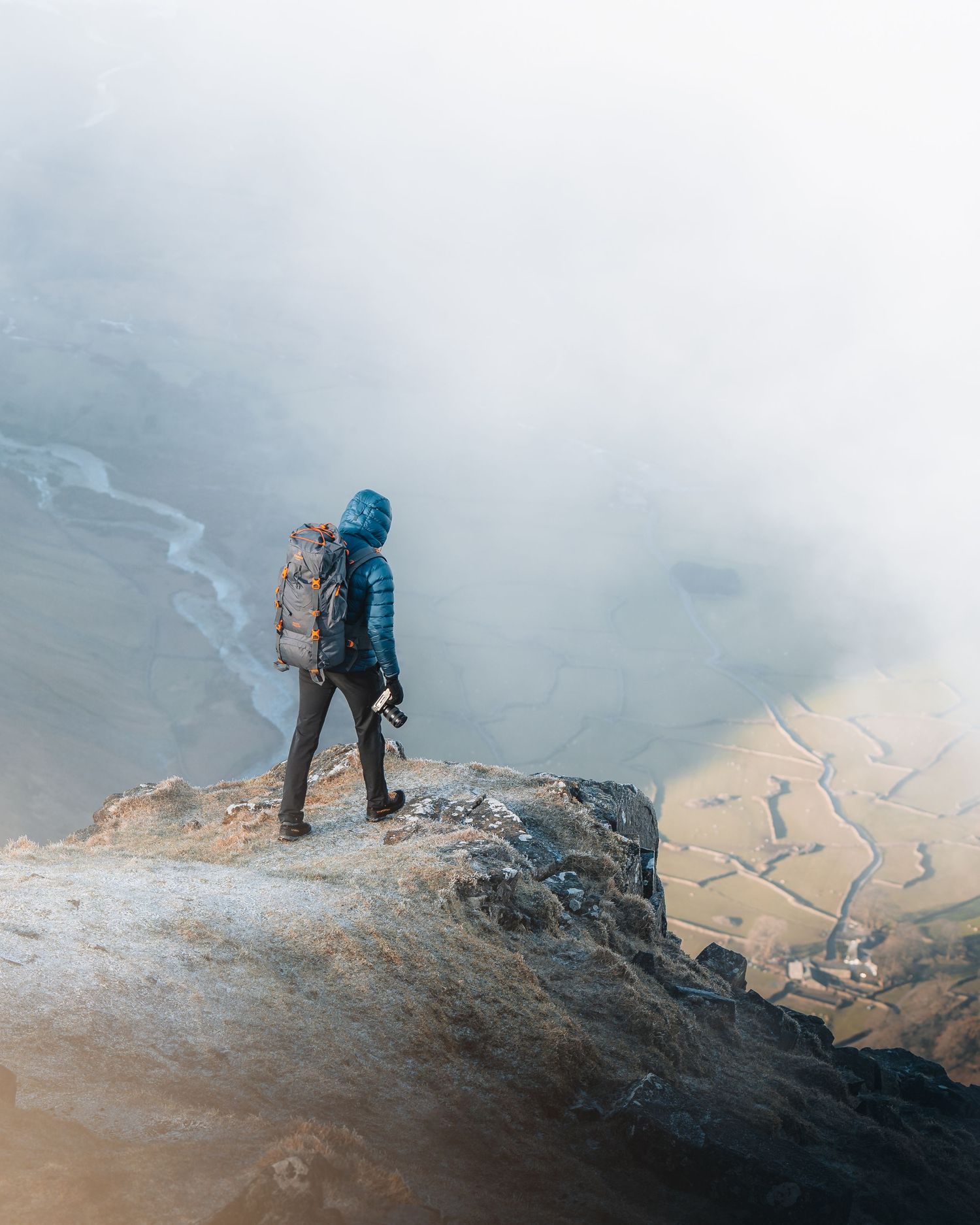 A hiker stands on a rocky cliff edge overlooking a misty valley with winding streams below during foggy conditions.
