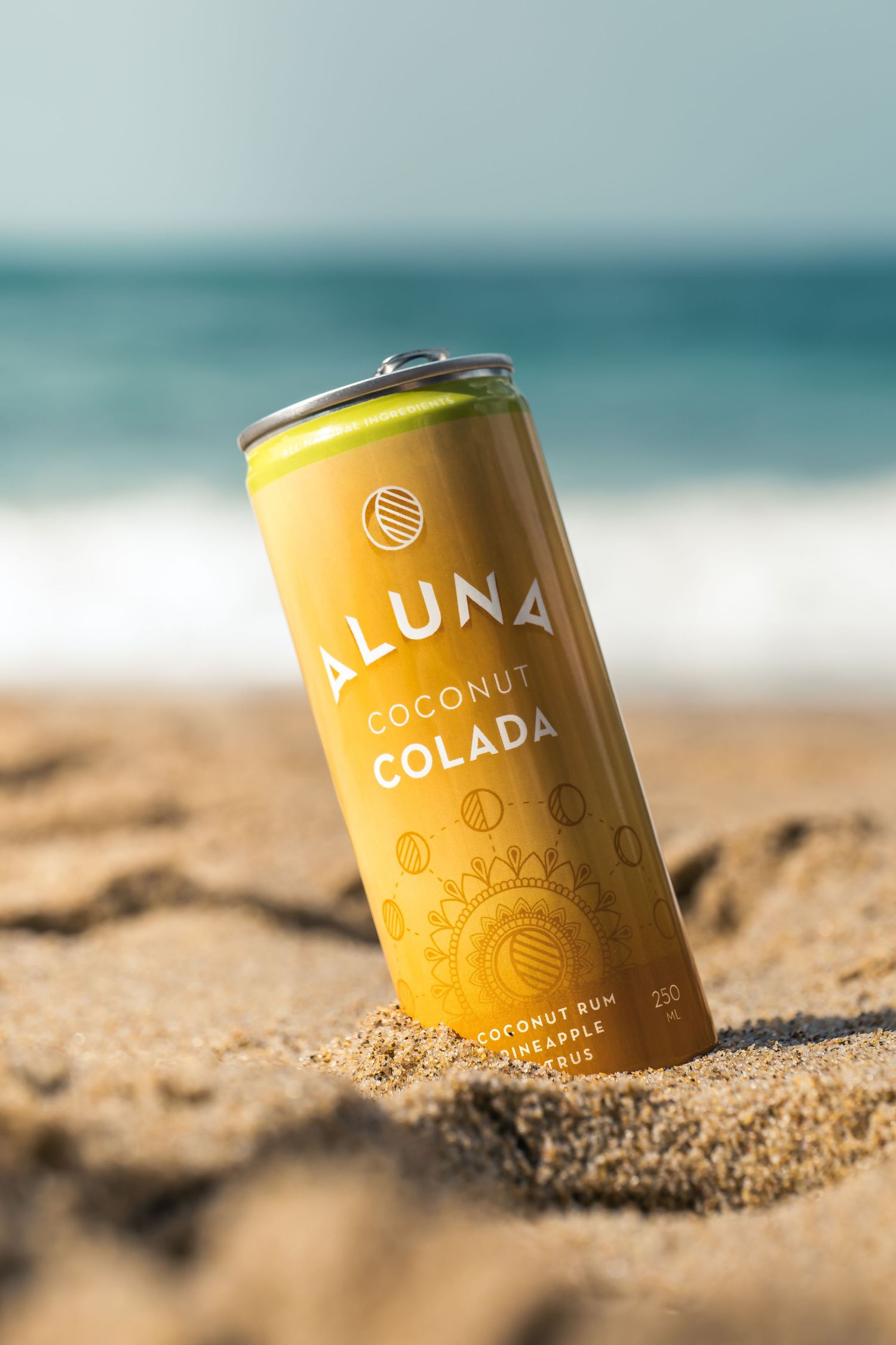 A yellow Coluna coconut drink can nestled in beach sand with ocean waves in the background.