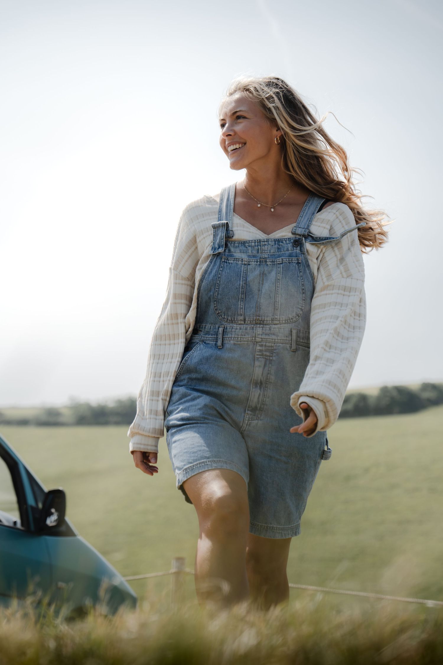 Woman in denim overalls and white sweater runs joyfully through a sunny meadow with long grass.
