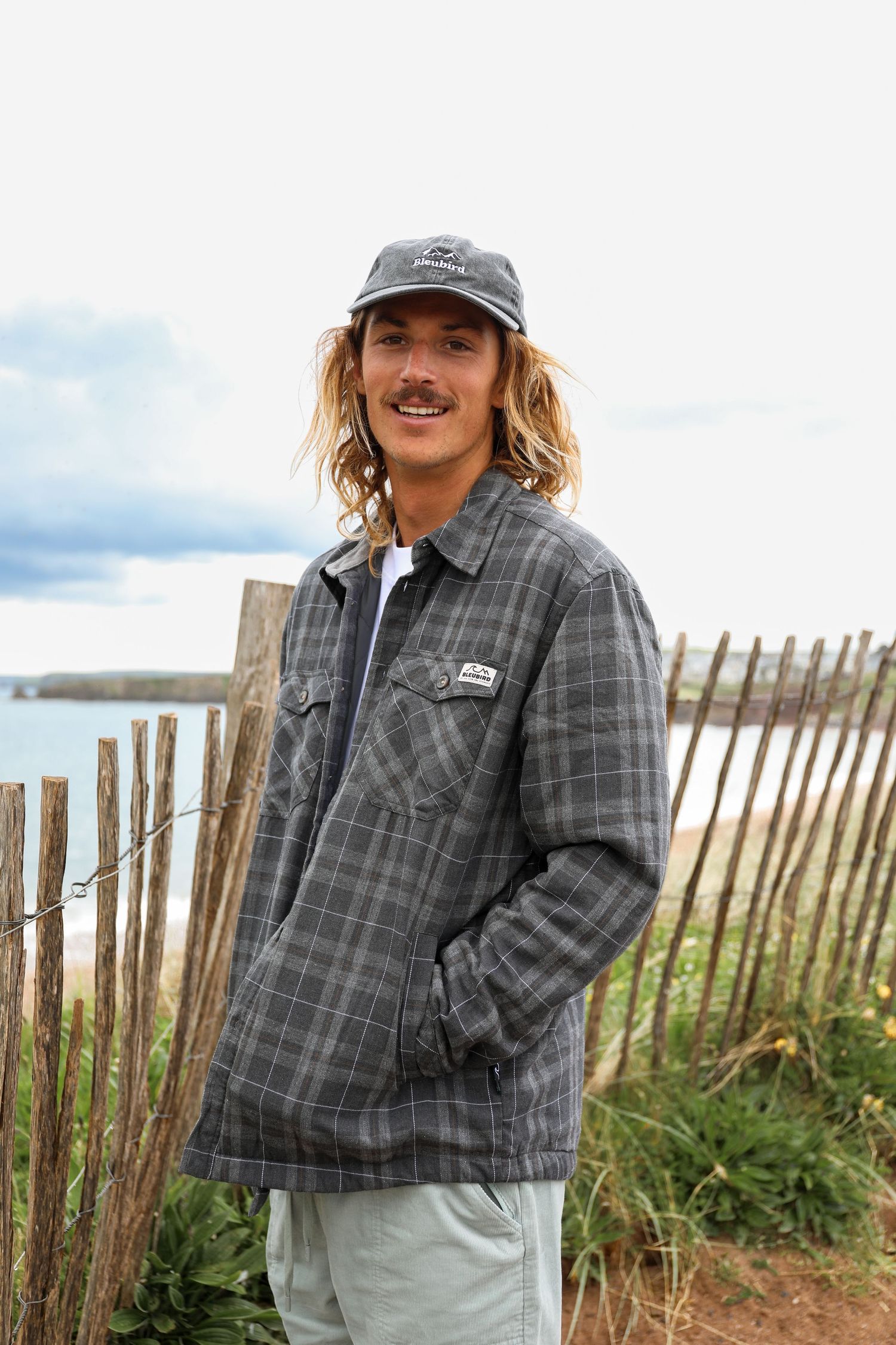 Surfer in a gray plaid flannel jacket and baseball cap stands by wooden fence at coastal viewpoint overlooking the ocean.