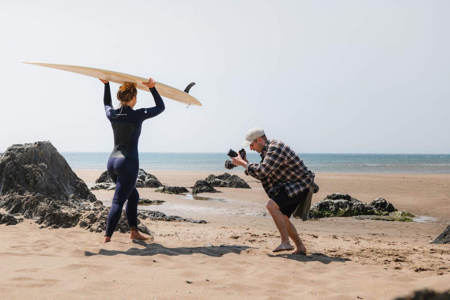 Two people engage playfully on a rocky beach, with one holding a surfboard and another photographing the scene.