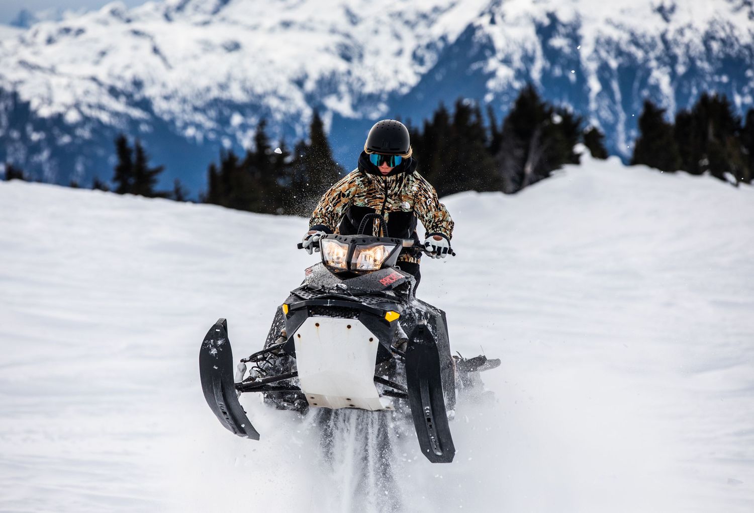 A snowmobiler catches air while jumping on snowy mountain terrain with snow-capped peaks in the background.